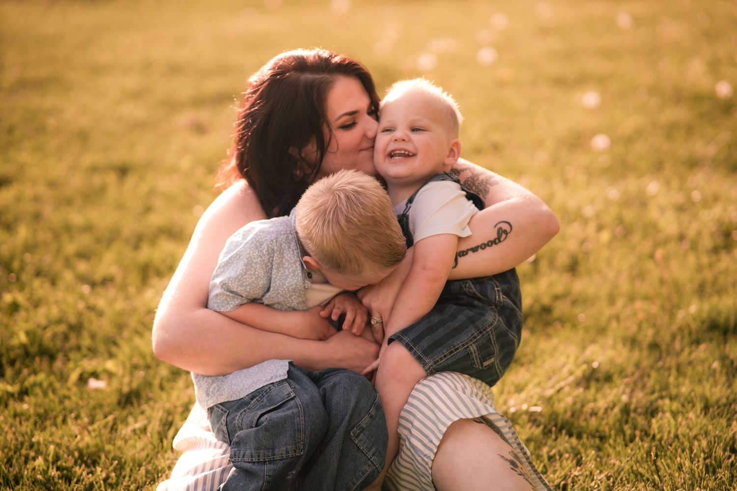 A mother sitting on green grass, hugging and kissing her two young boys in warm golden sunlight.