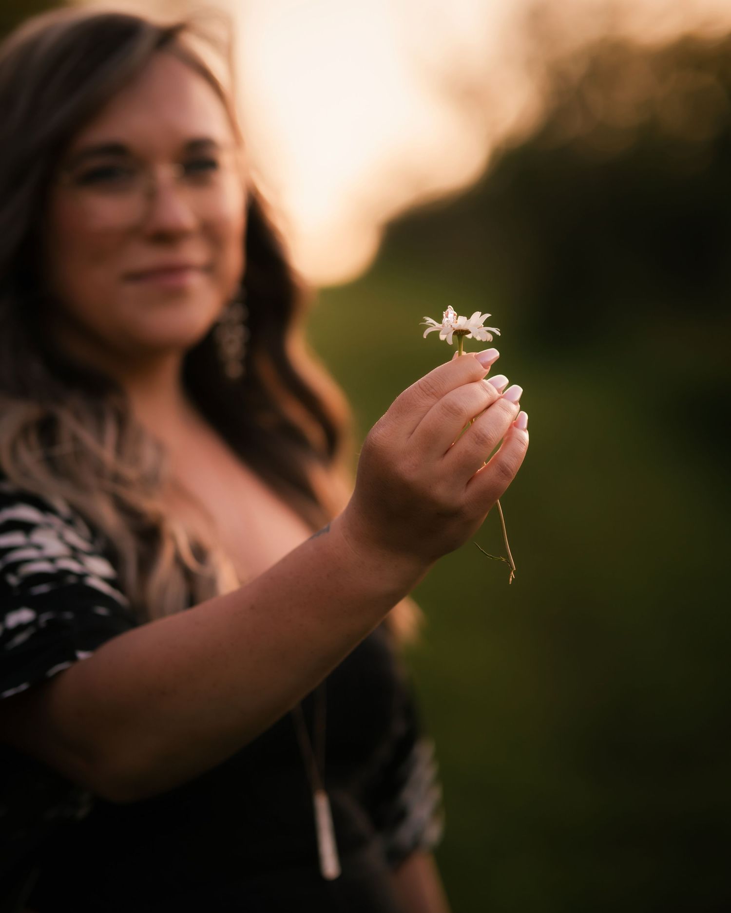 Woman holding a small white wildflower toward camera at golden hour with blurred green background.