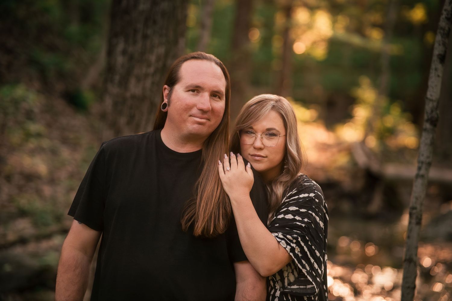 Couple posing together in a sunlit forest, woman resting her head on mans shoulder, wearing casual attire.