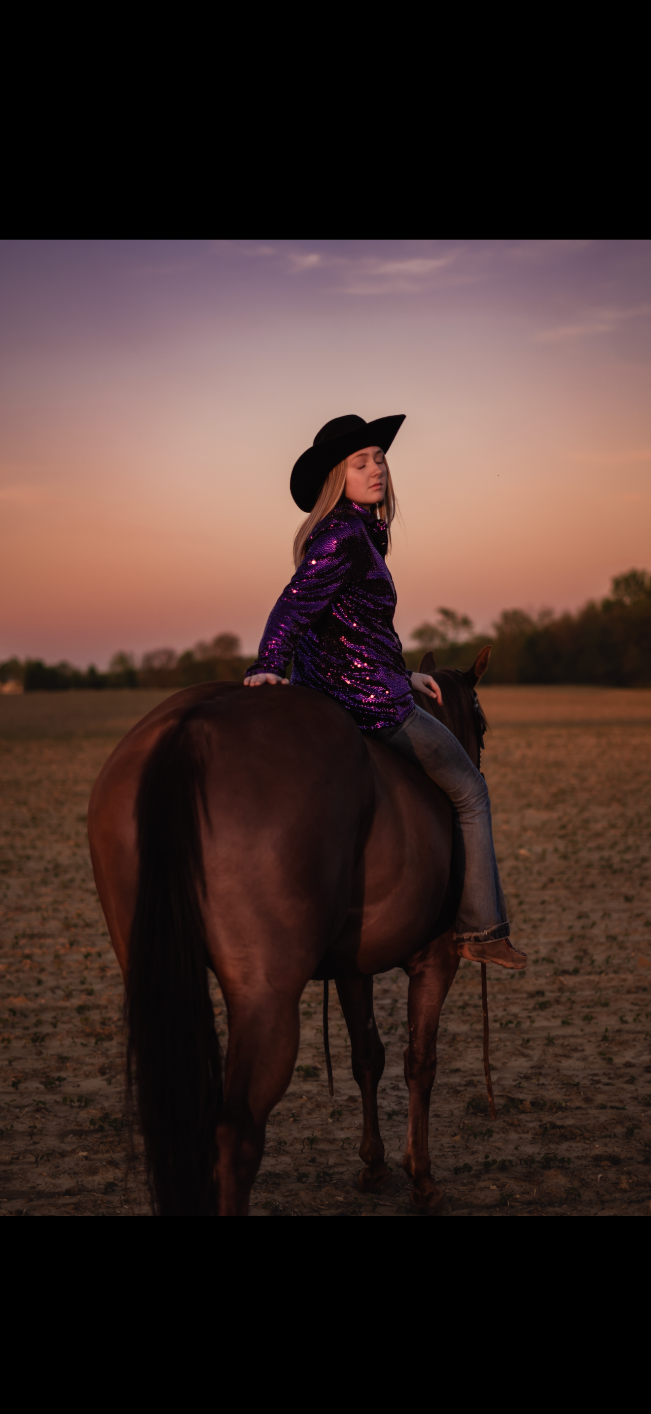 A rider in a sparkly purple jacket and black hat sits atop a brown horse at sunset.