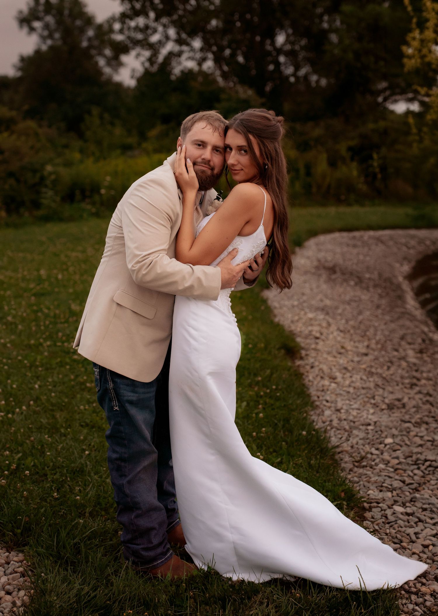 Bride in white gown and groom in tan blazer embrace outdoors on gravel path surrounded by lush greenery.