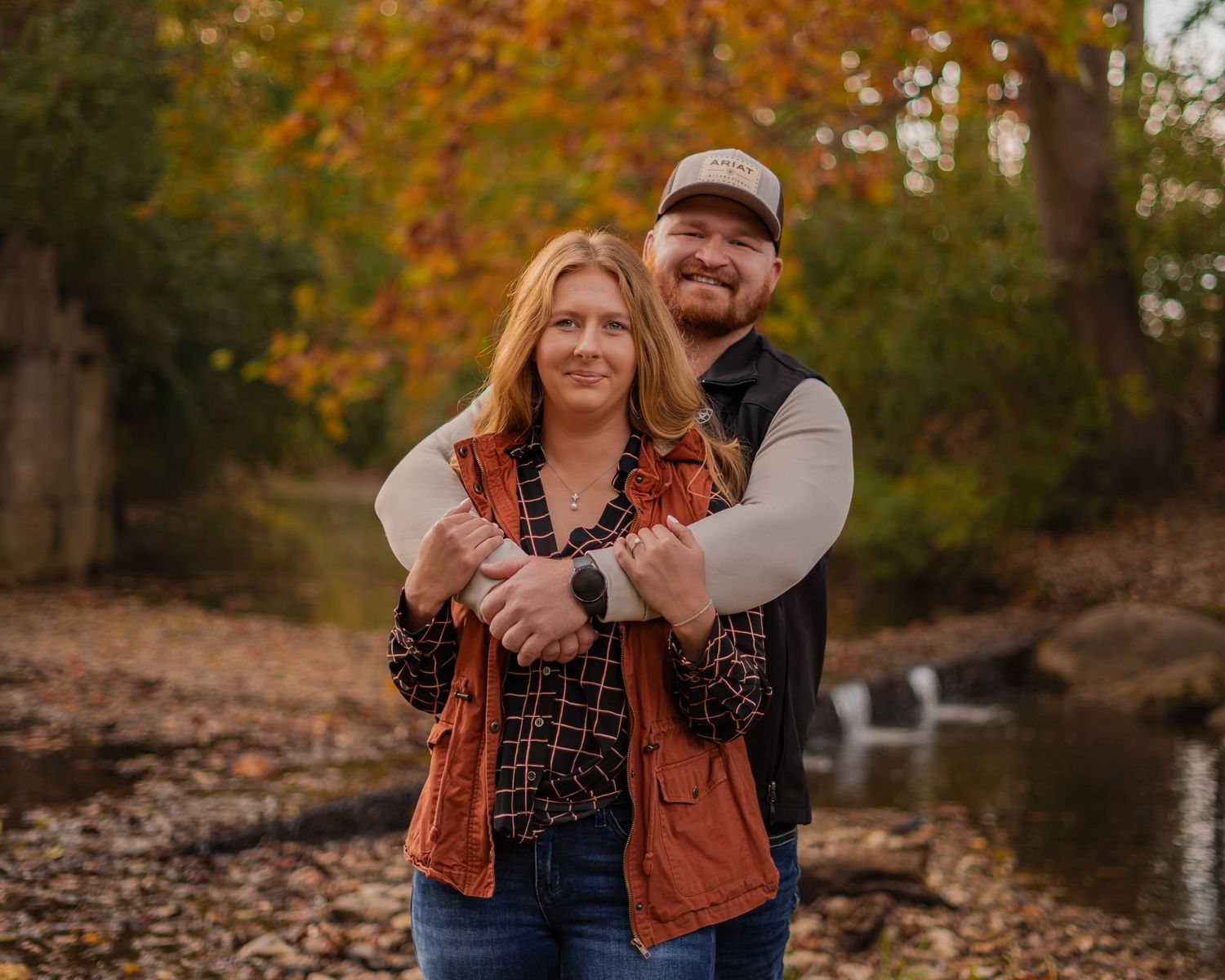 Couple embraces in a romantic autumn setting with fallen leaves and trees.