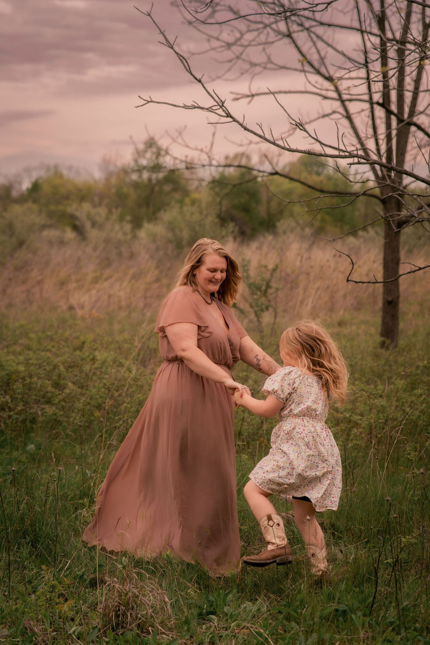 A mother and young daughter dance together in a field at sunset, wearing flowy dresses near a bare tree.