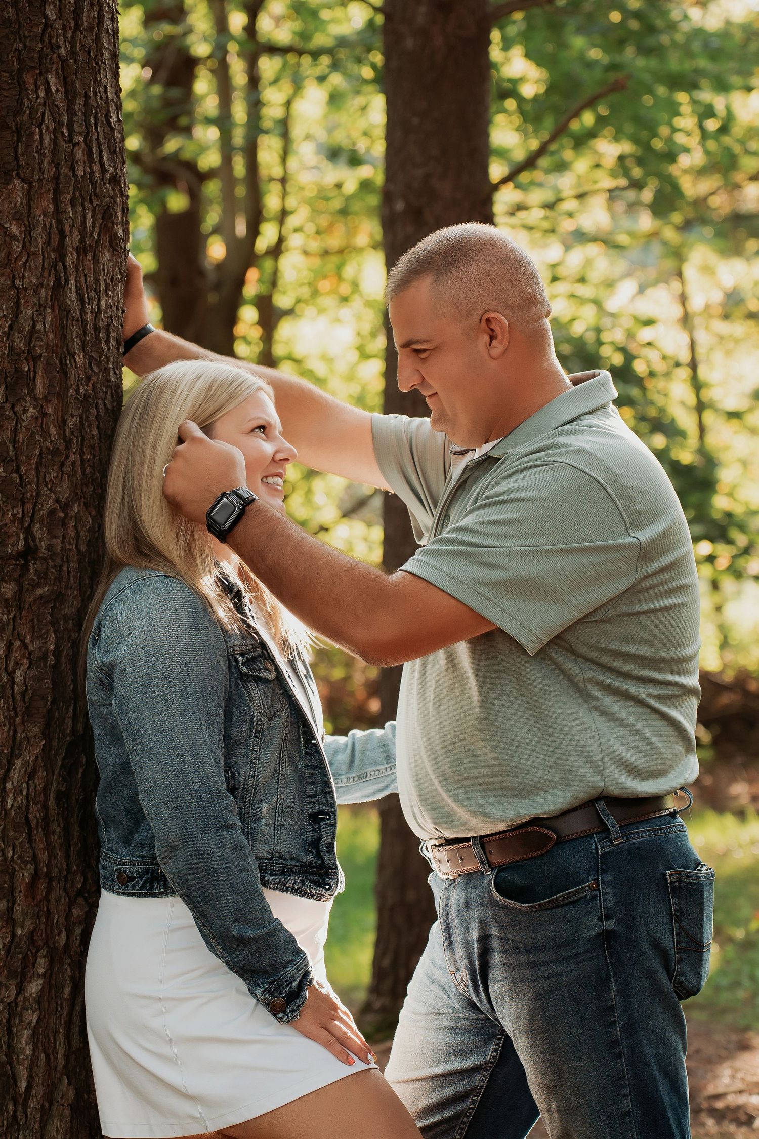 A couple shares a tender moment near a tree in a sunlit forest, the man gently touching the woman's face.