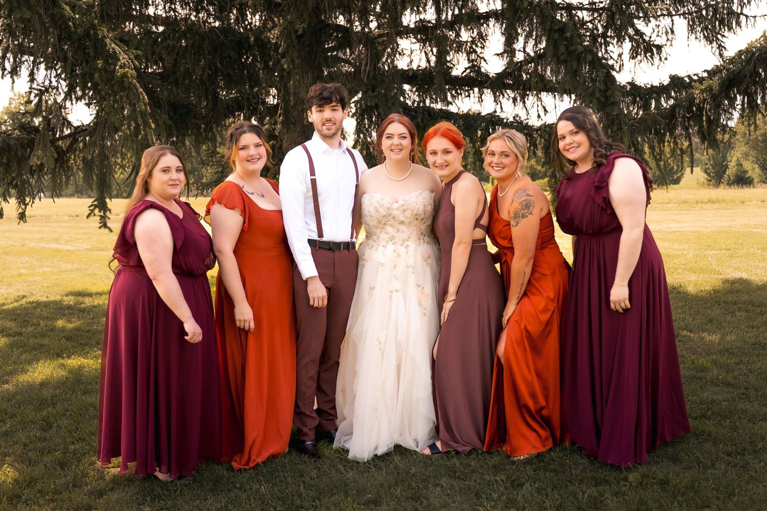 Wedding party posing outdoors, bride in white gown with groom in suspenders surrounded by bridesmaids in burgundy and rust gowns.