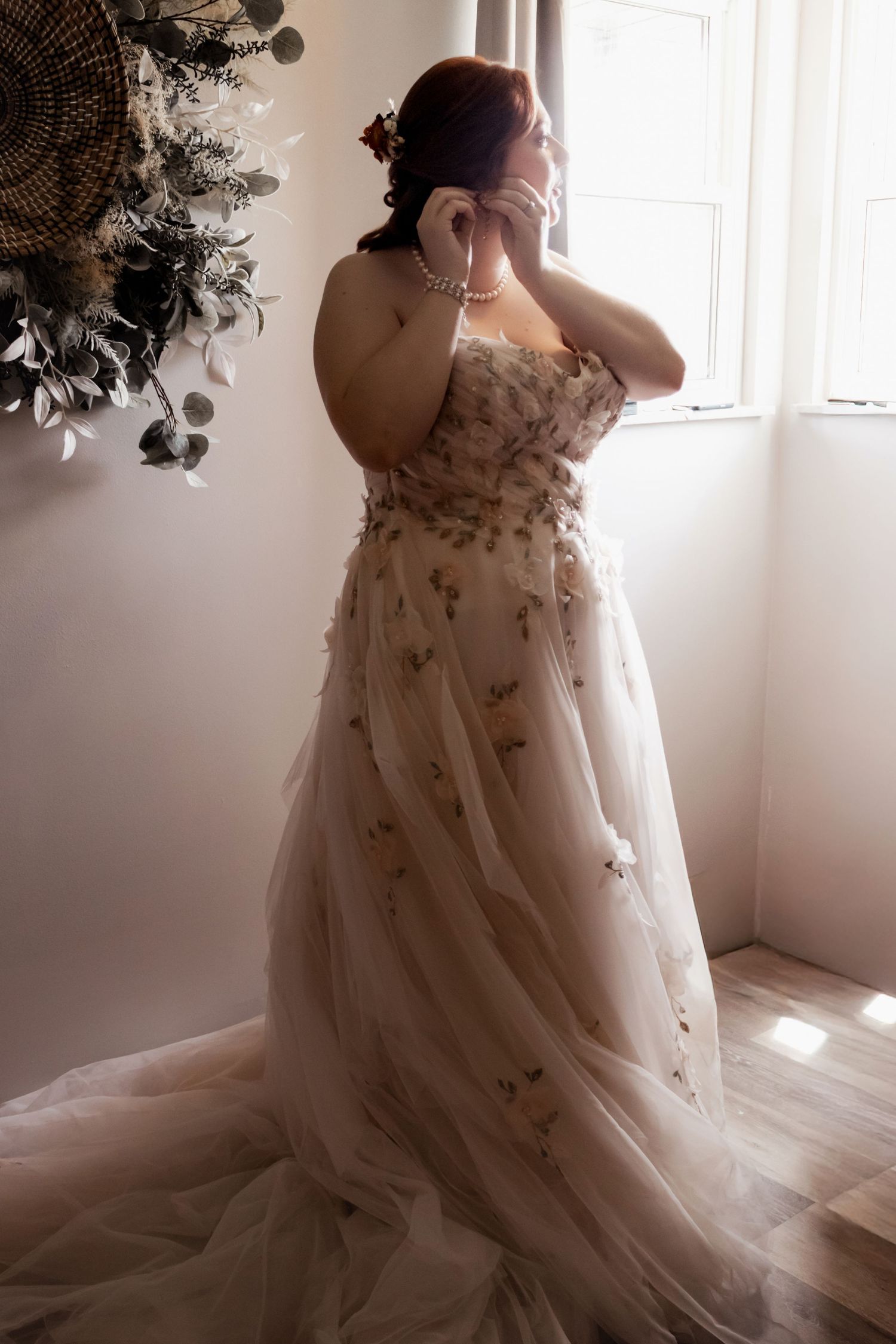 Bride in floral tulle gown with pearl necklace stands by window in soft natural light.