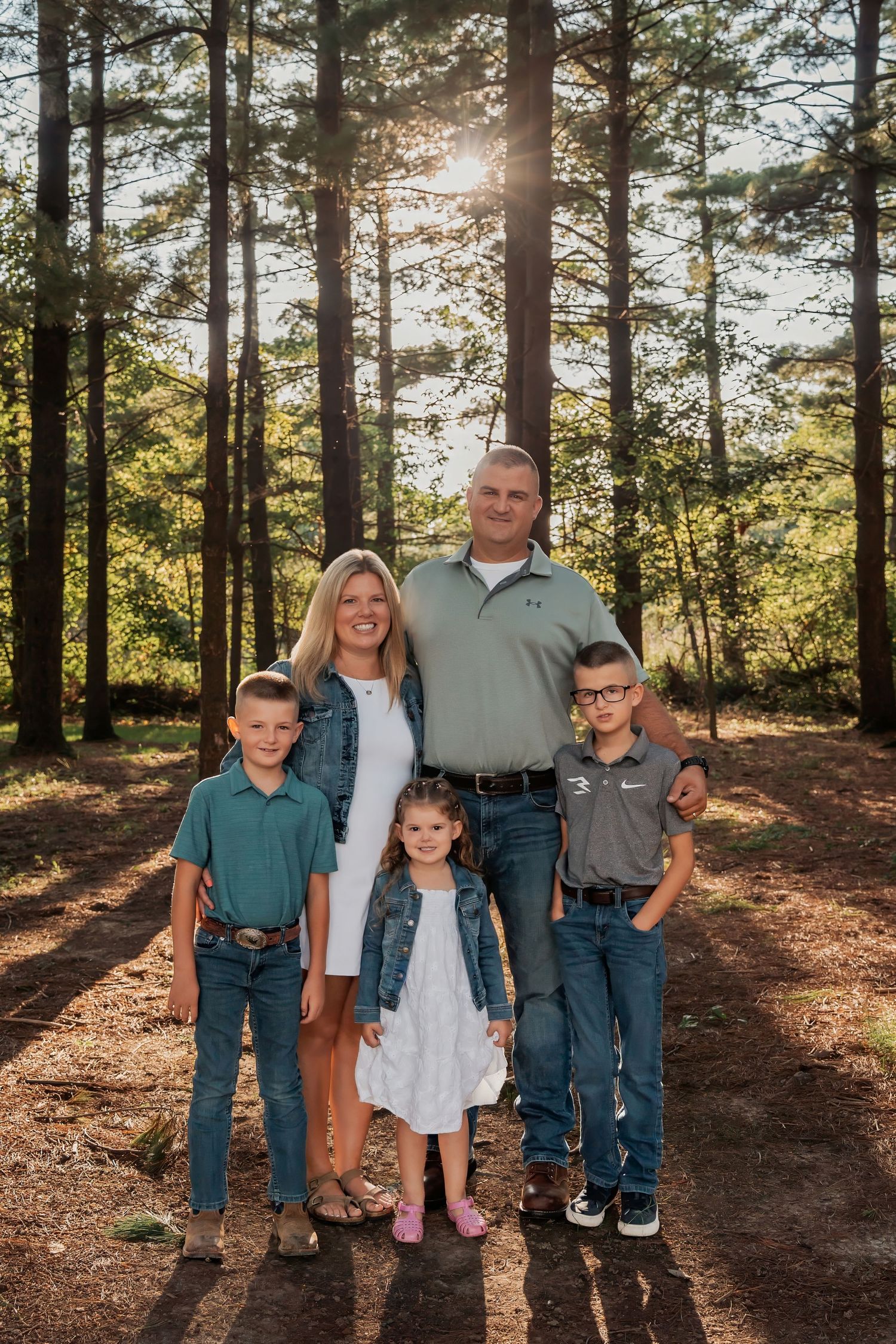 Family of five posing together in a sunlit pine forest, parents with three young children dressed in casual coordinated outfits.