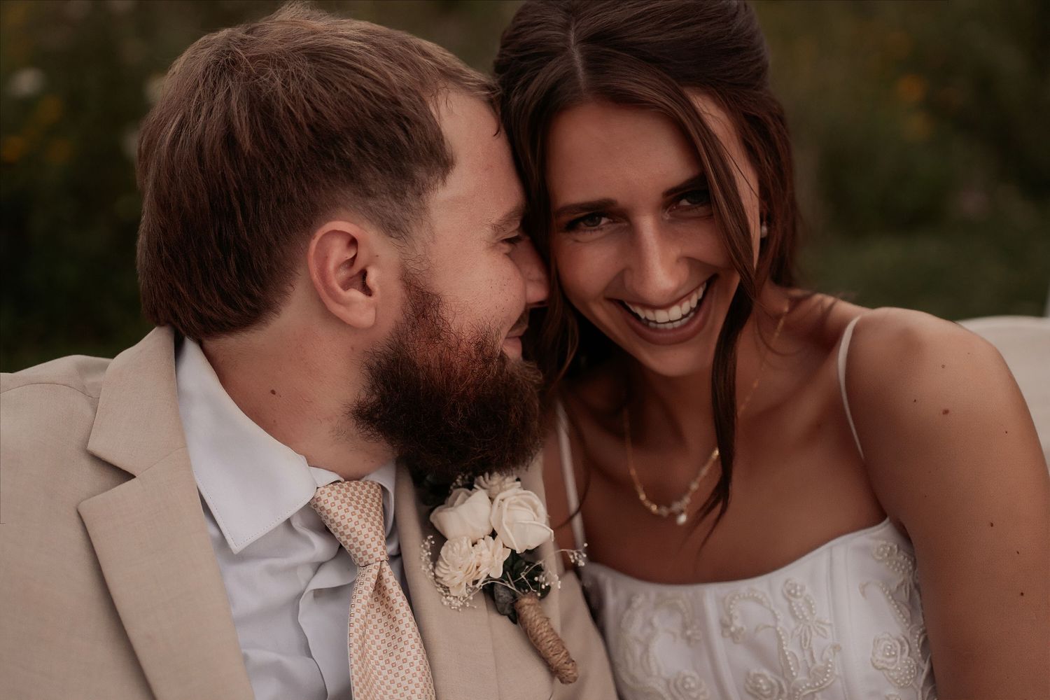 Joyful bride and bearded groom in tan suit share a tender, laughing moment outdoors on their wedding day.