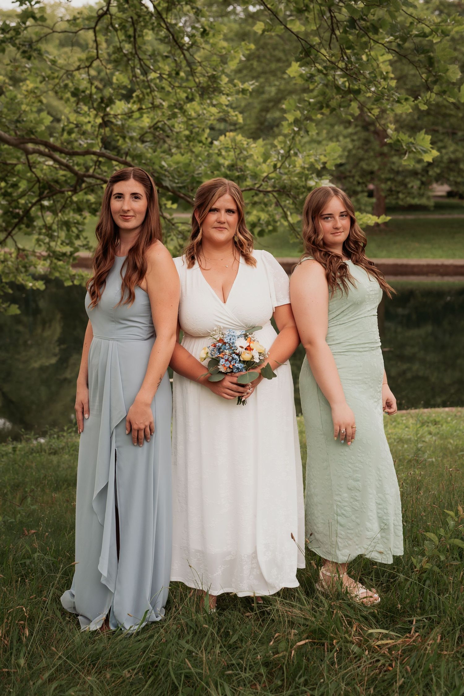 Bride in white dress holding bouquet poses with two bridesmaids in sage green gowns outdoors near trees.