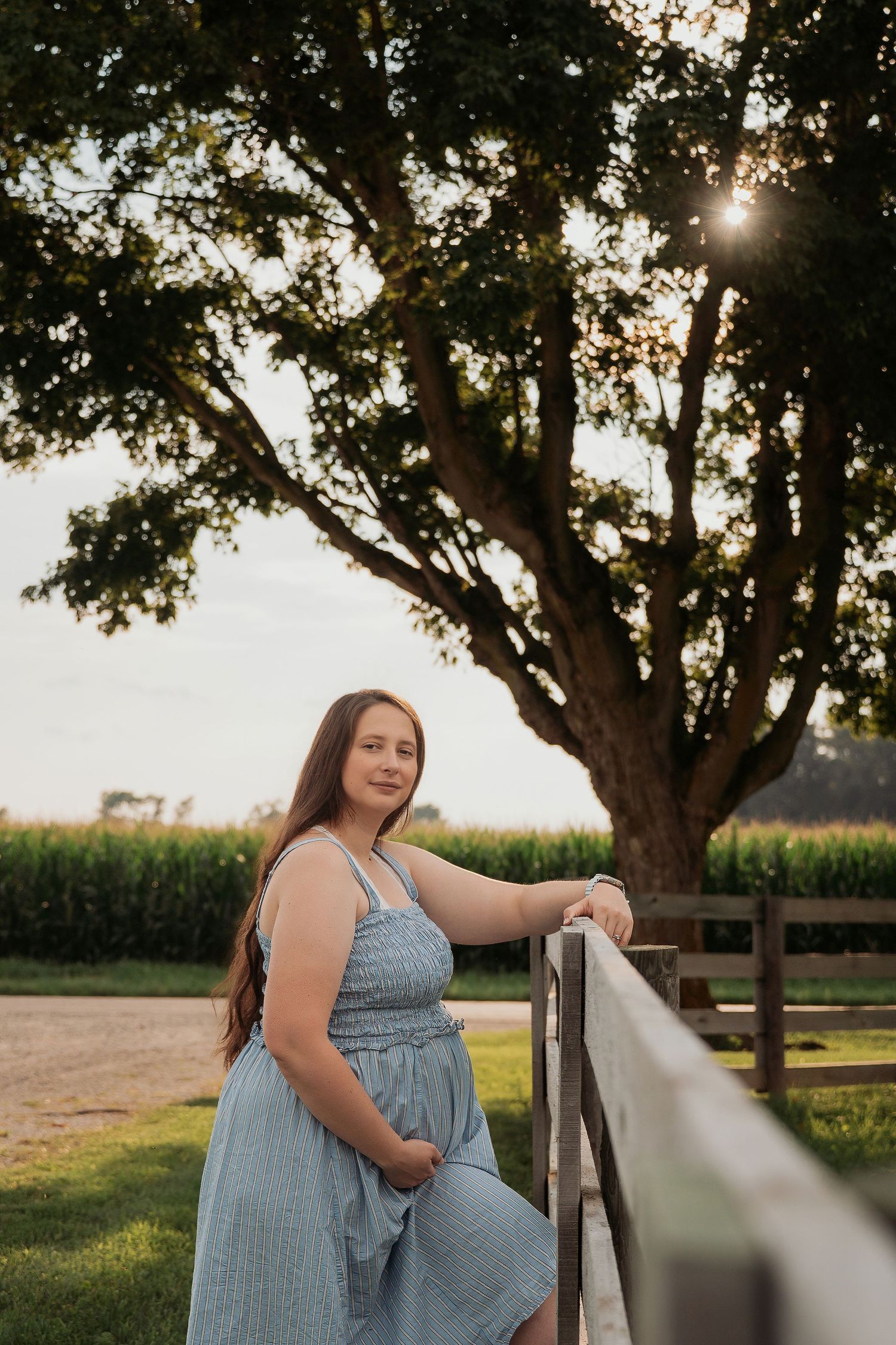 Young woman in blue sundress leaning on wooden fence at golden hour with large tree and cornfield backdrop.