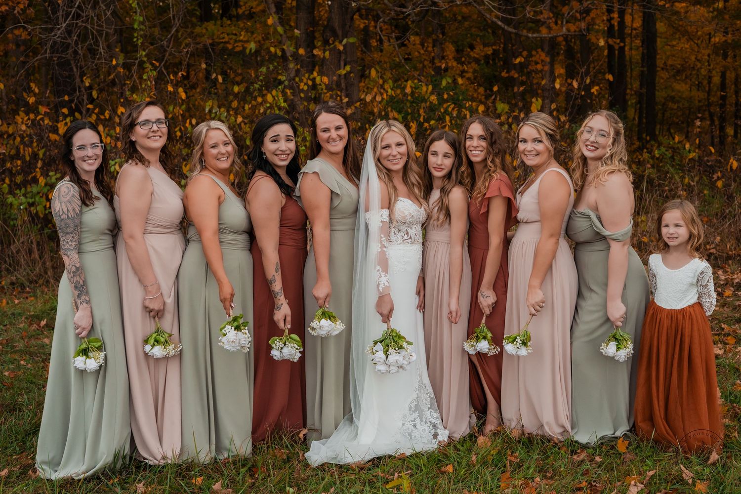 A bridal party in coordinated pink and green dresses poses together in an autumn setting.