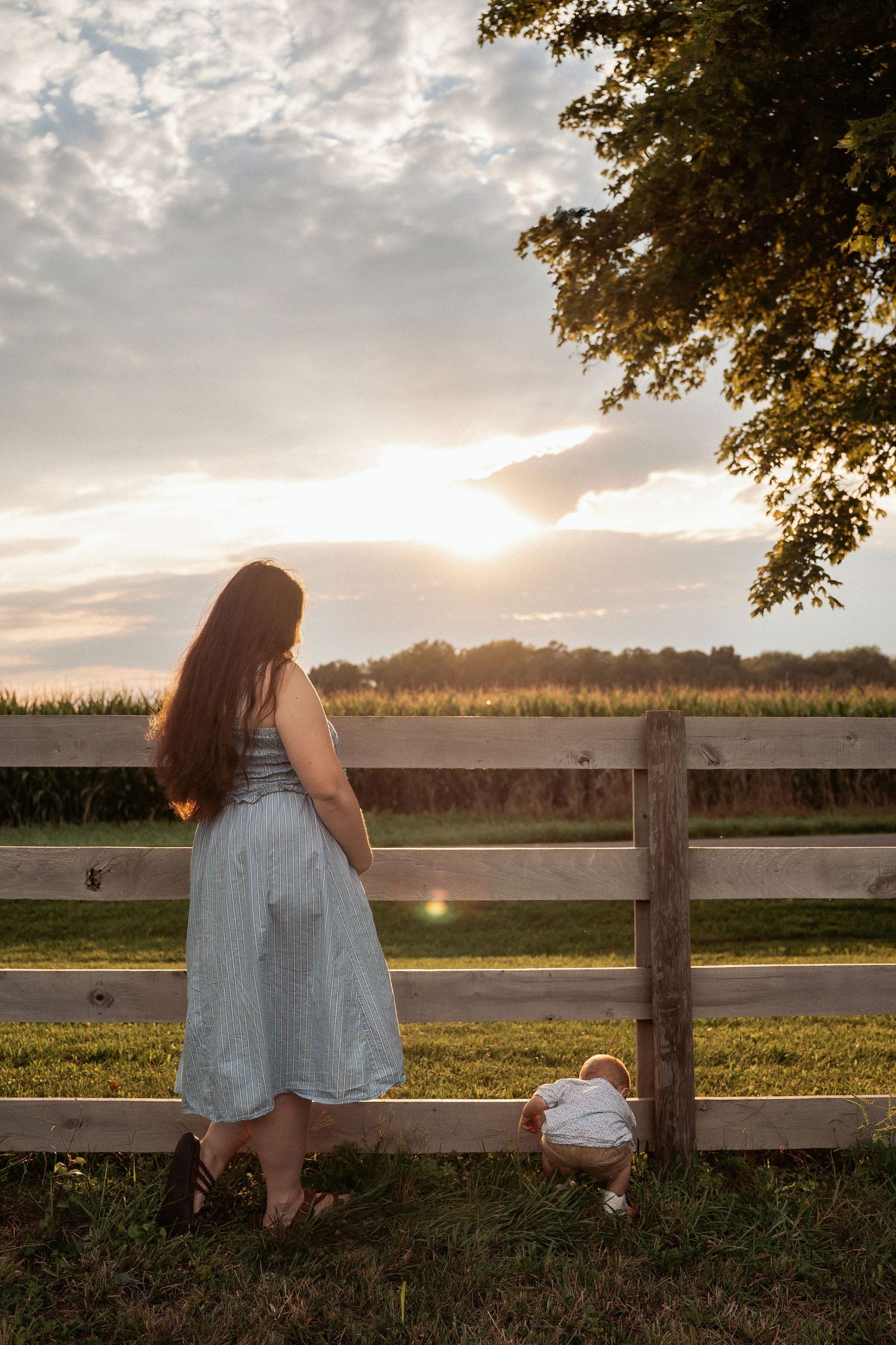 Woman in blue dress standing by wooden fence with toddler at sunset near cornfield.
