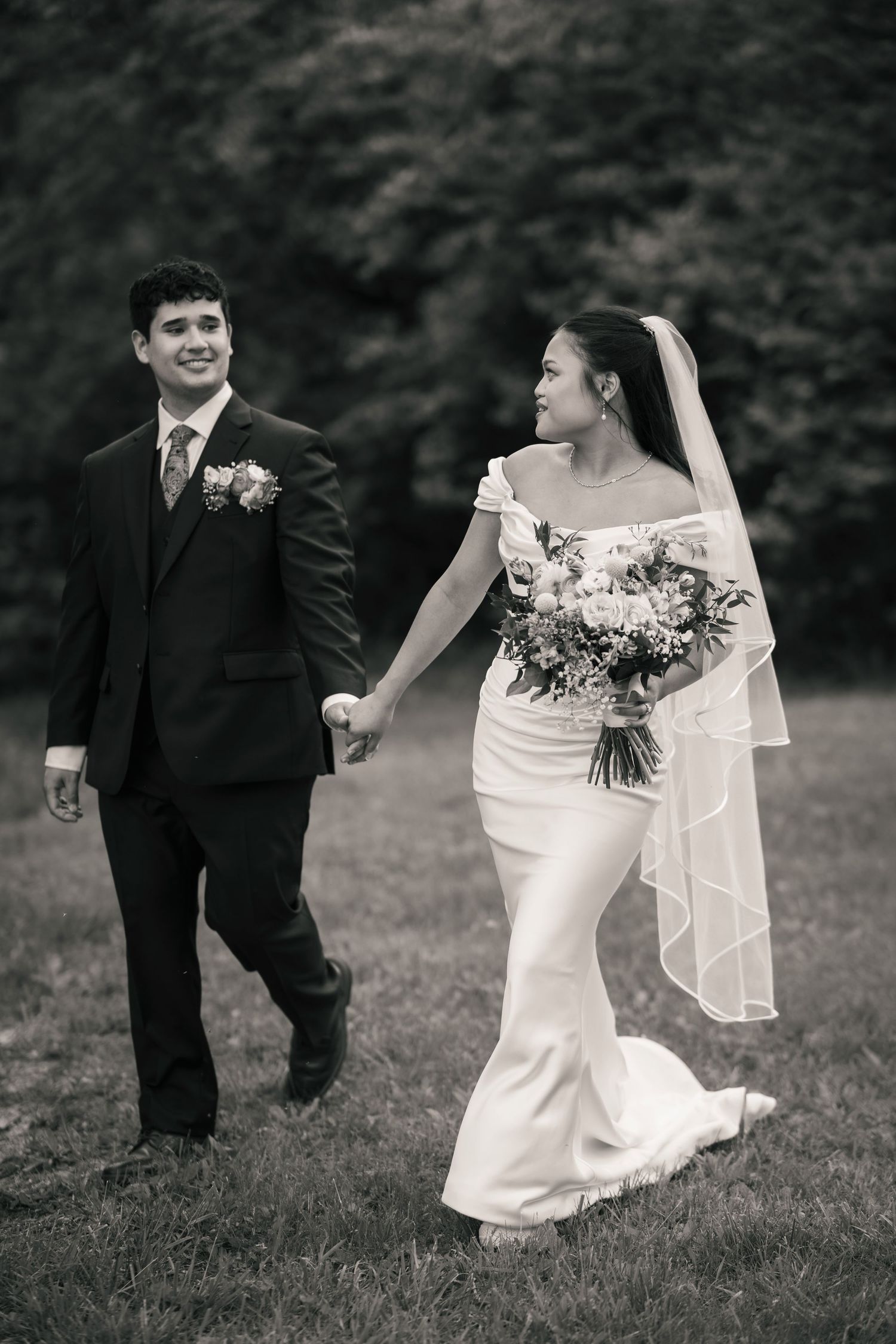 Black and white photo of a smiling bride and groom holding hands while walking outdoors on their wedding day.