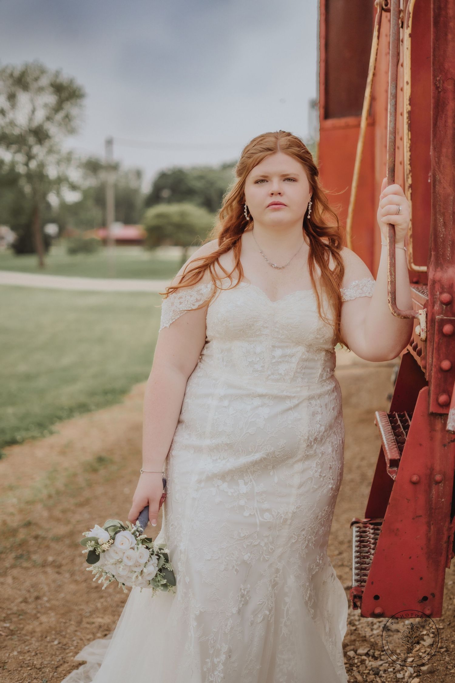Bride in lace wedding dress poses against red rustic structure holding white bouquet.