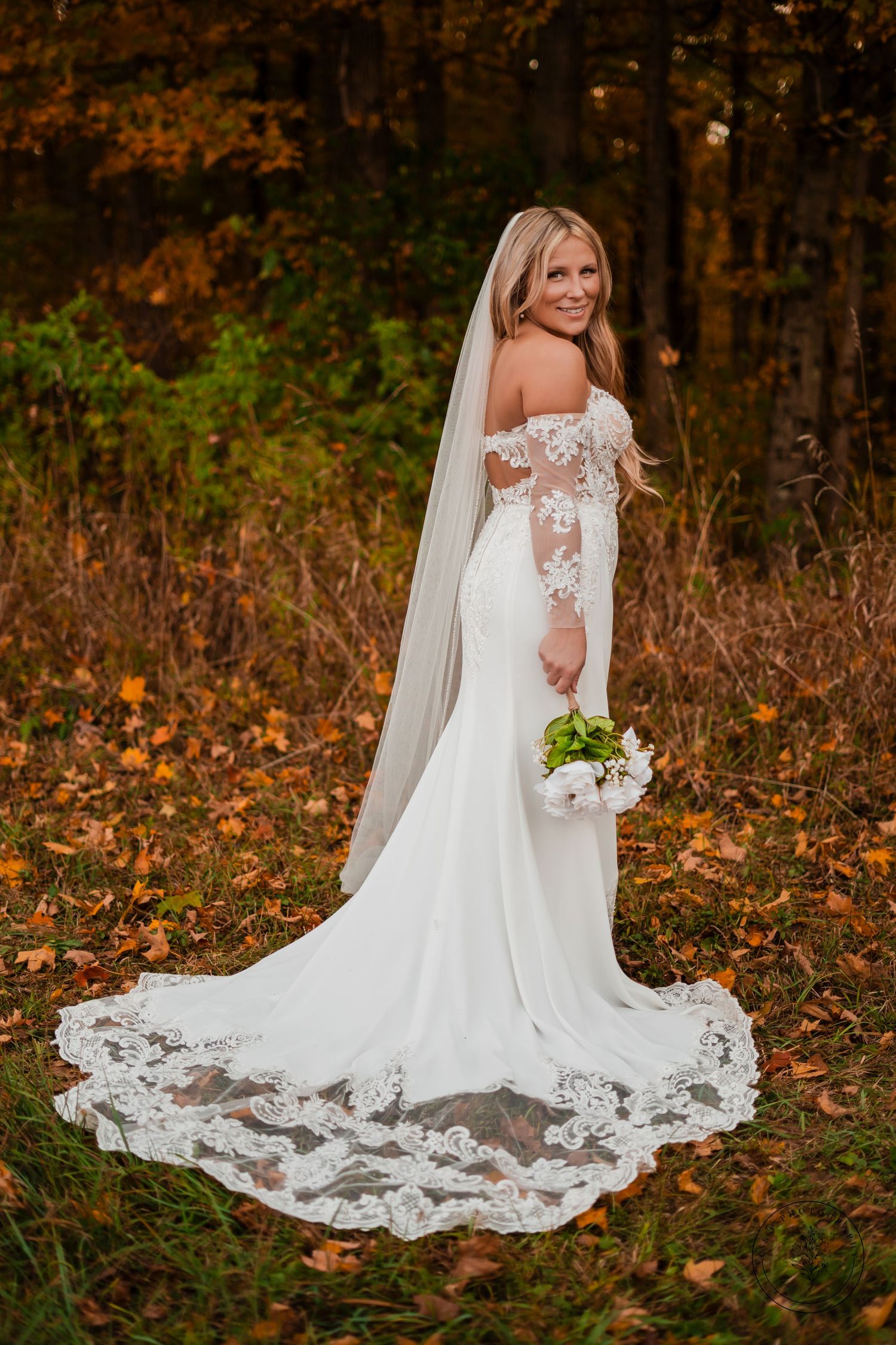 Elegant bride in lace off-shoulder wedding gown with long train poses in autumn foliage.