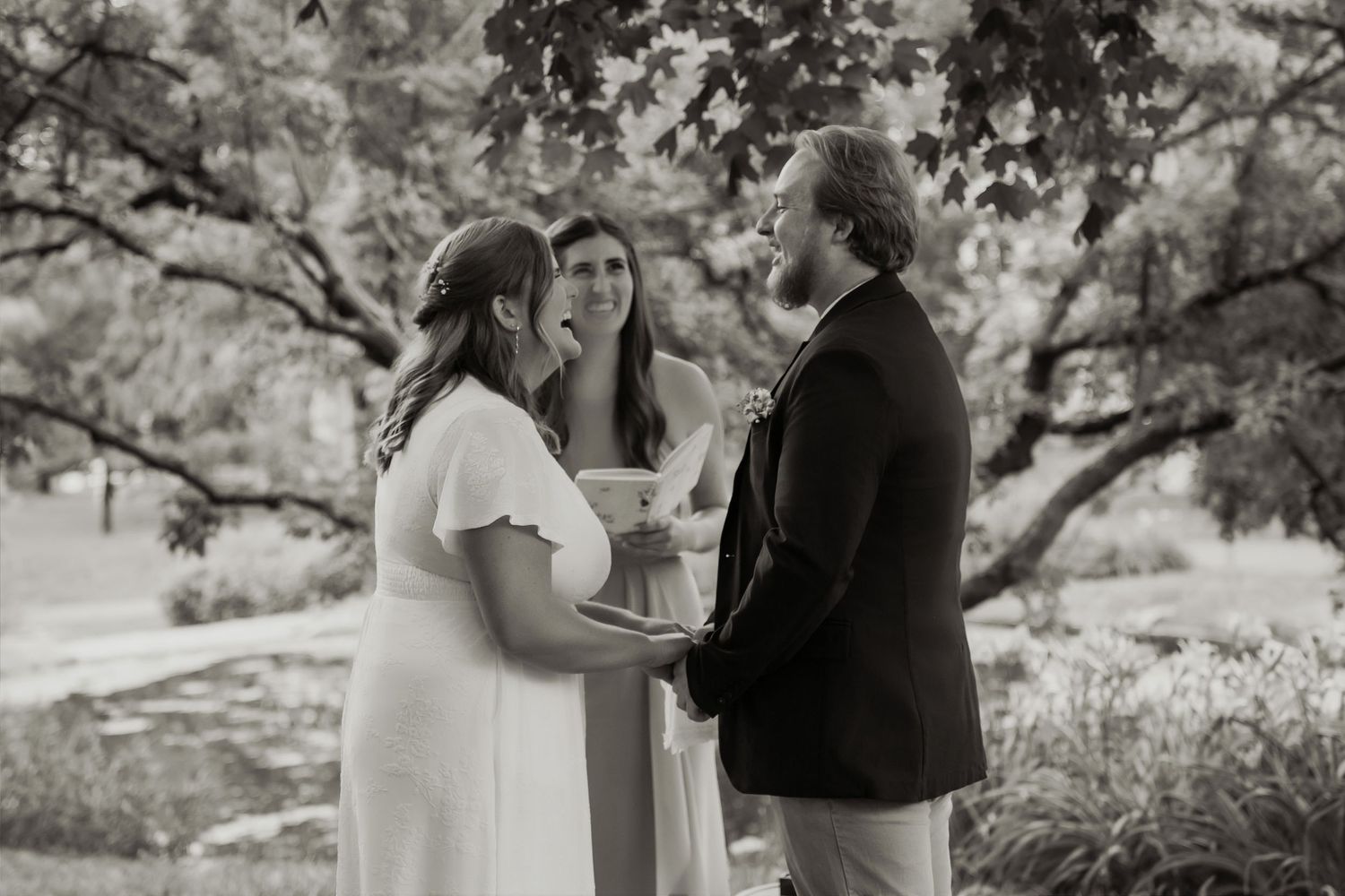 Black and white outdoor wedding ceremony with bride in white dress and groom in dark suit holding hands before officiant.