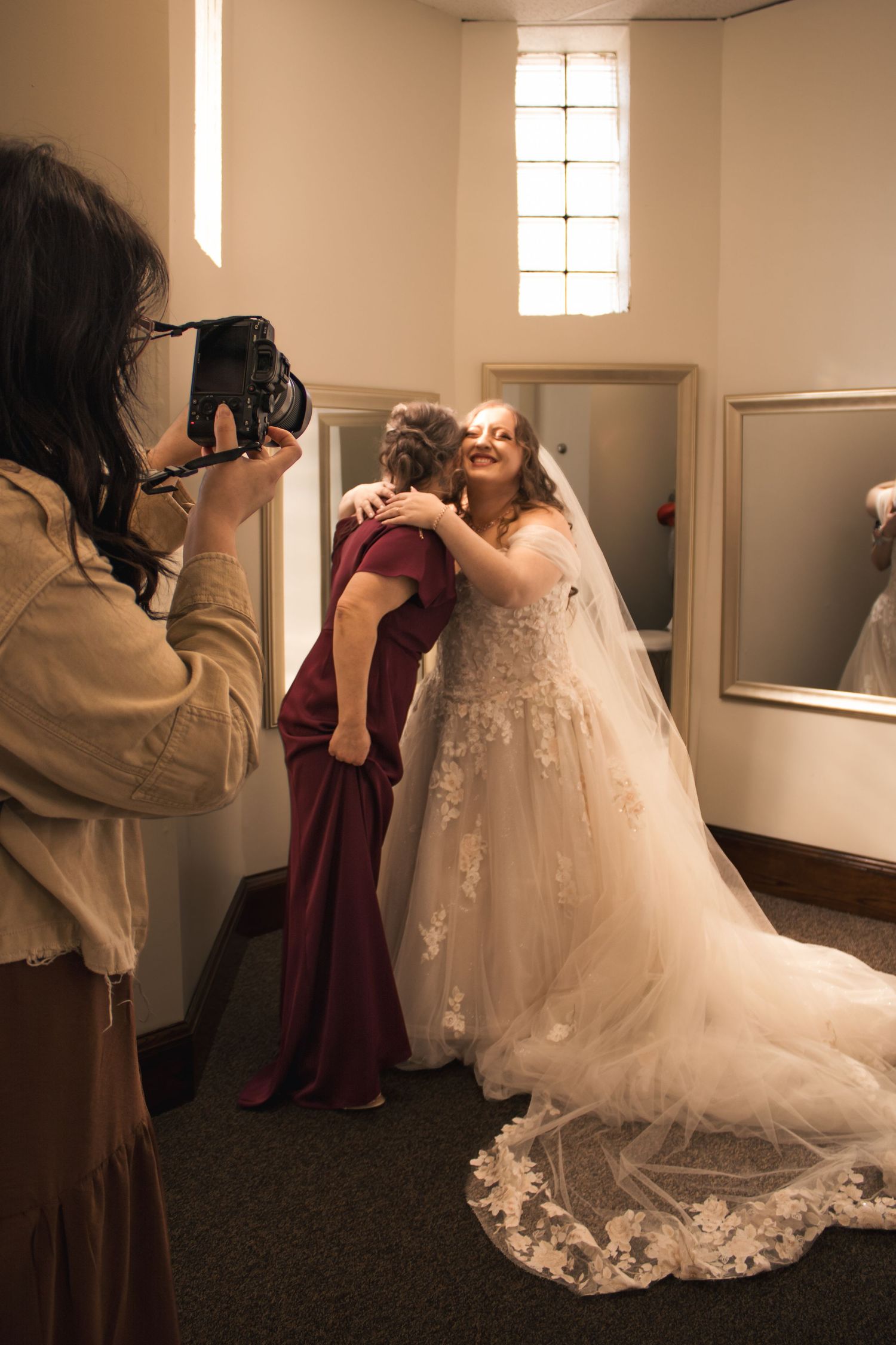 Bride in lace gown hugging woman in burgundy dress while photographer captures the emotional moment in bridal suite.