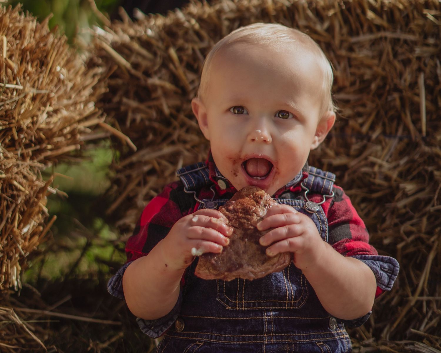 Baby in plaid outfit plays with autumn leaves while sitting outdoors.