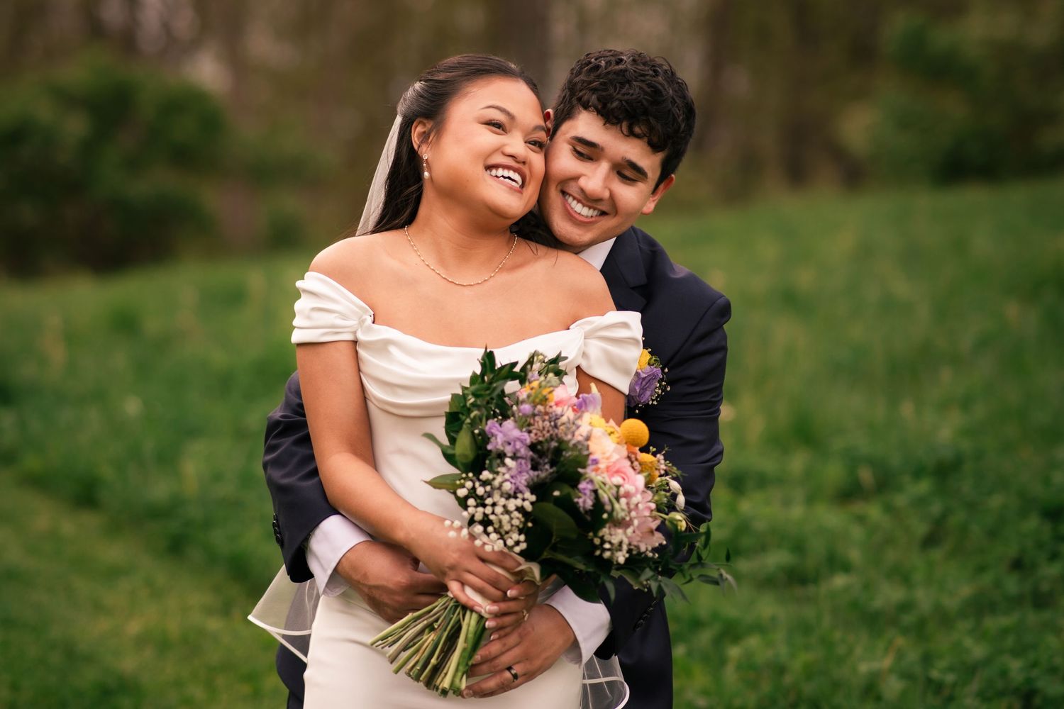 Happy couple in wedding attire embracing outdoors, bride holding colorful bouquet in white off-shoulder gown.