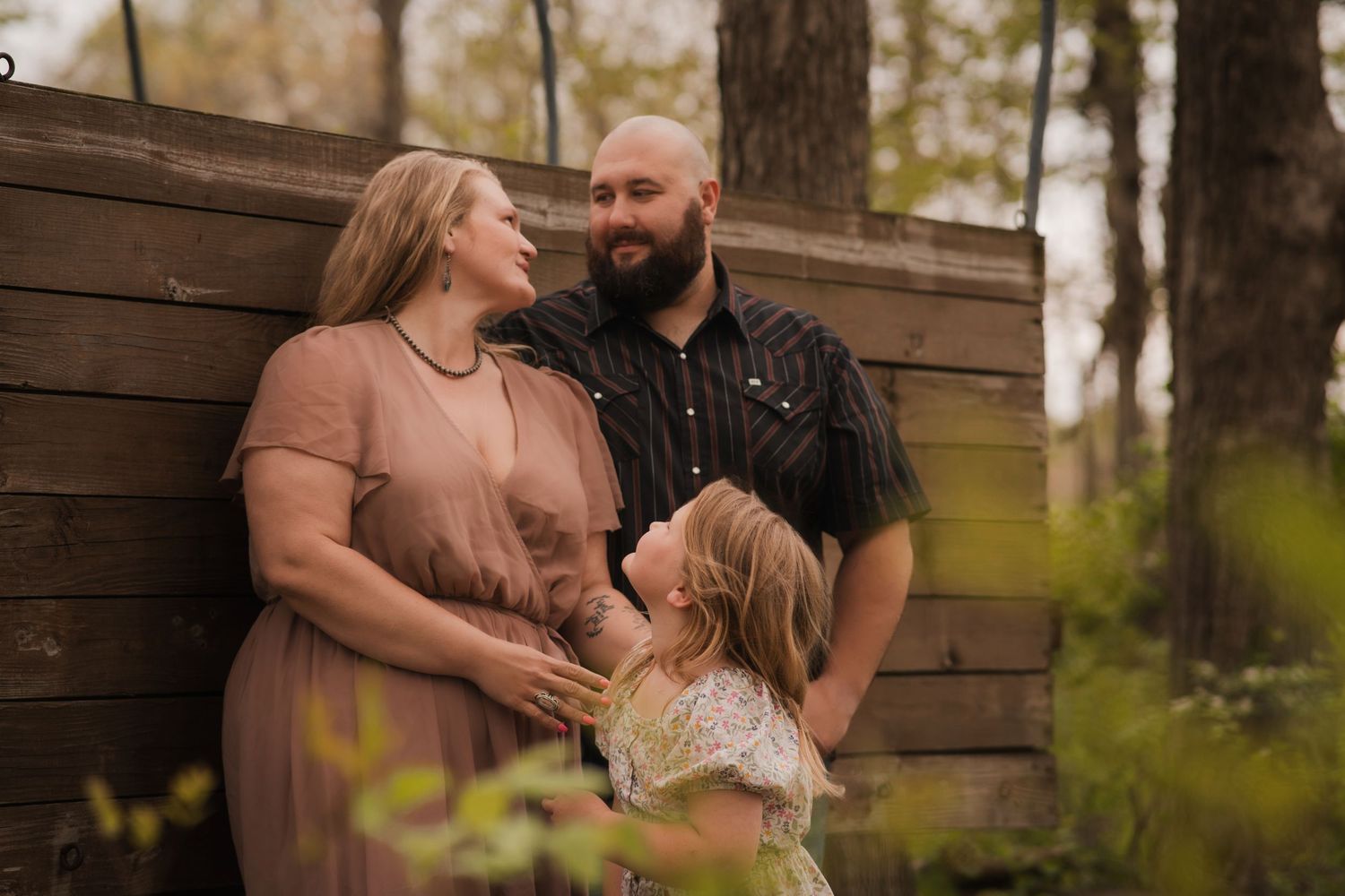 A happy family of three poses outdoors near a wooden fence, with parents smiling at their young daughter in a woodland setting.