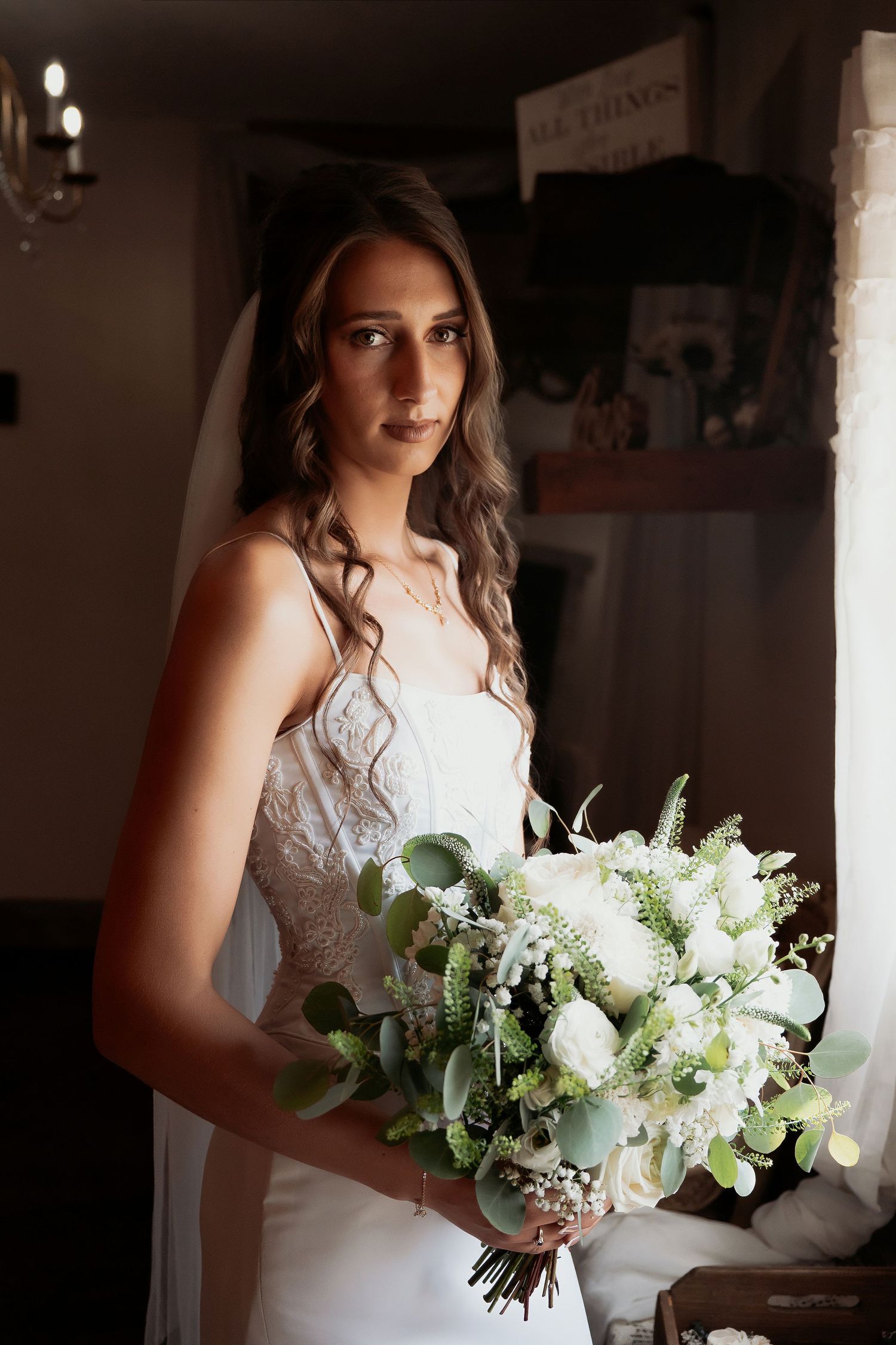 Bride in white lace gown holding elegant white floral bouquet with greenery, standing by window in soft natural light.