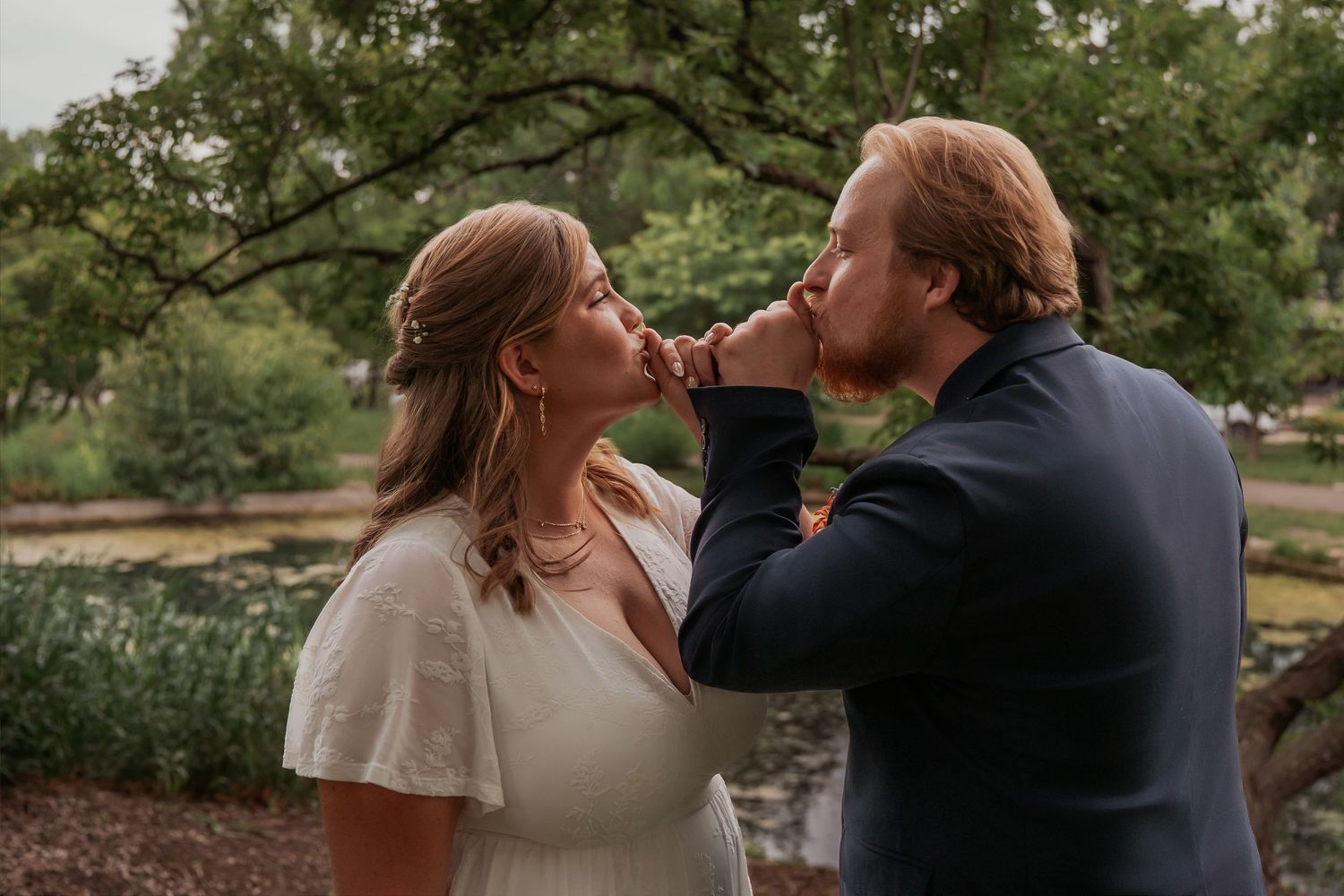 A bride and groom share a tender kiss outdoors near a pond surrounded by lush green trees at their wedding.