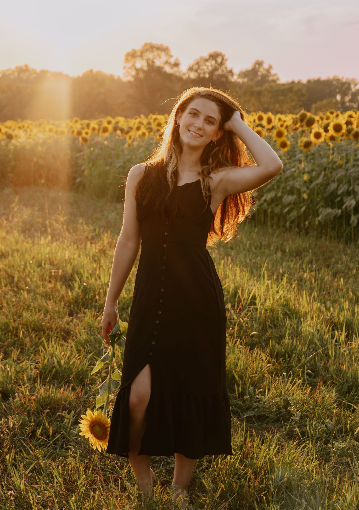 Woman in black dress poses in sunflower field during golden sunset.