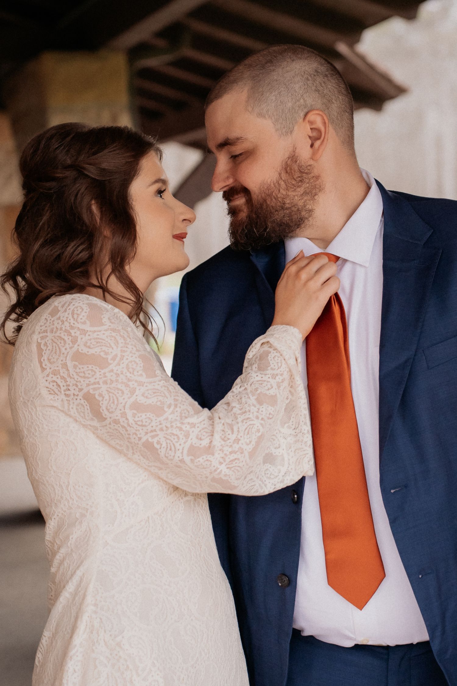 Bride in lace dress adjusting groom's orange tie, sharing a romantic moment in a wedding portrait.