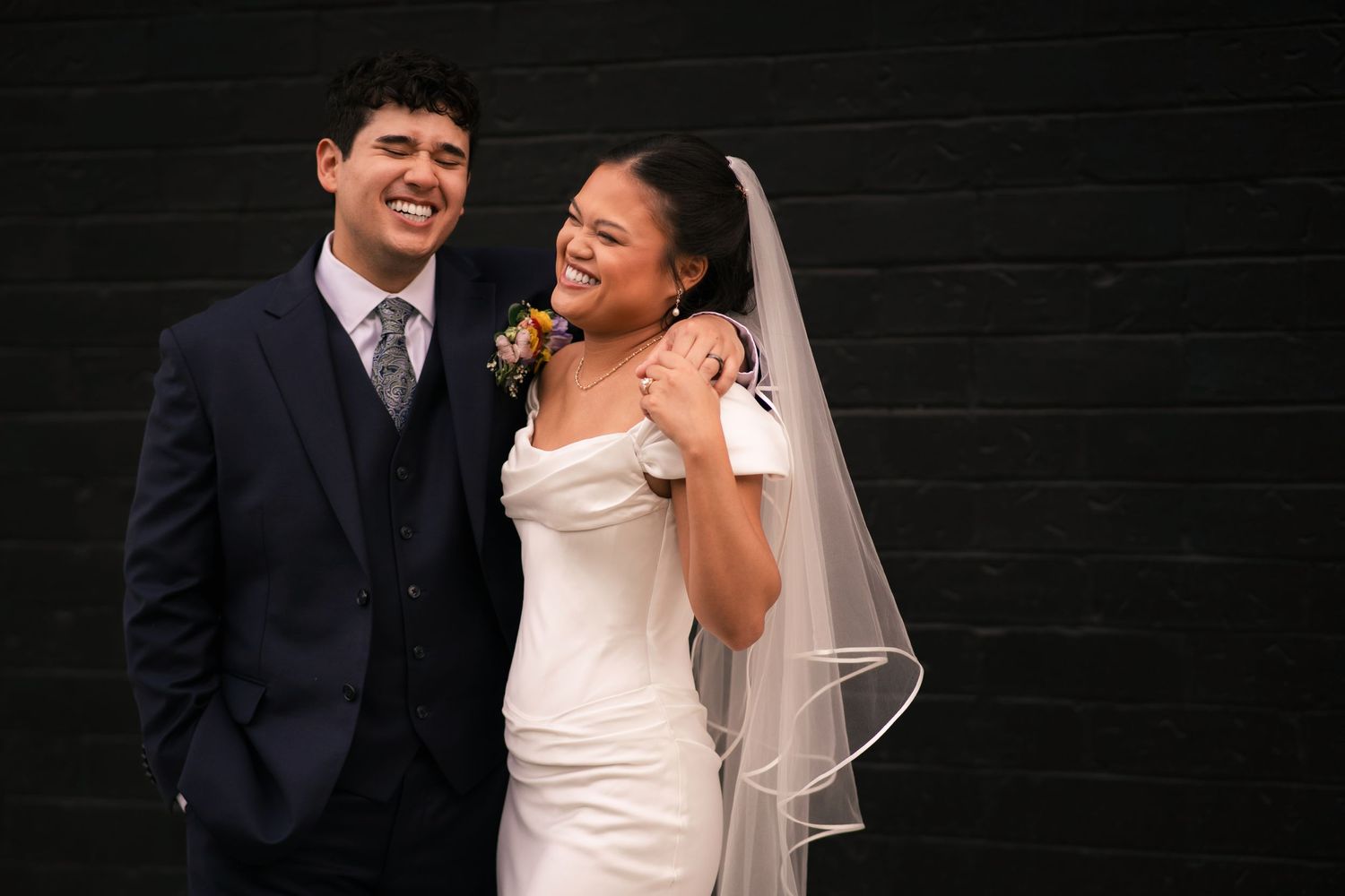 Smiling bride in white gown and veil poses with groom in navy suit against black brick wall.