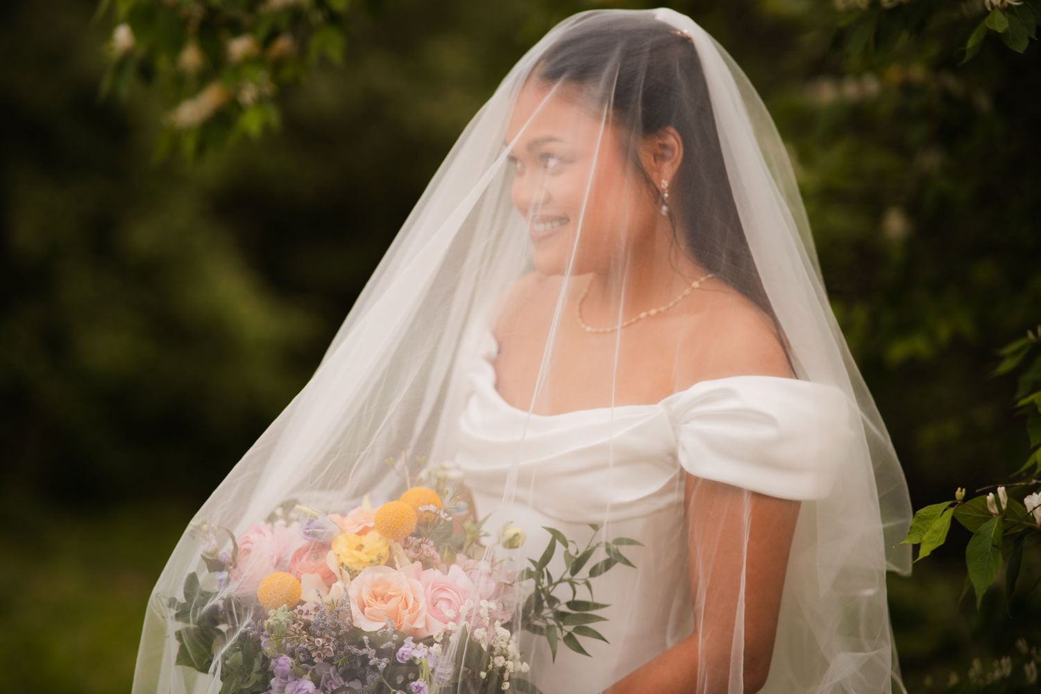 Bride in off-shoulder white gown holds colorful bouquet under flowing veil, surrounded by lush greenery.
