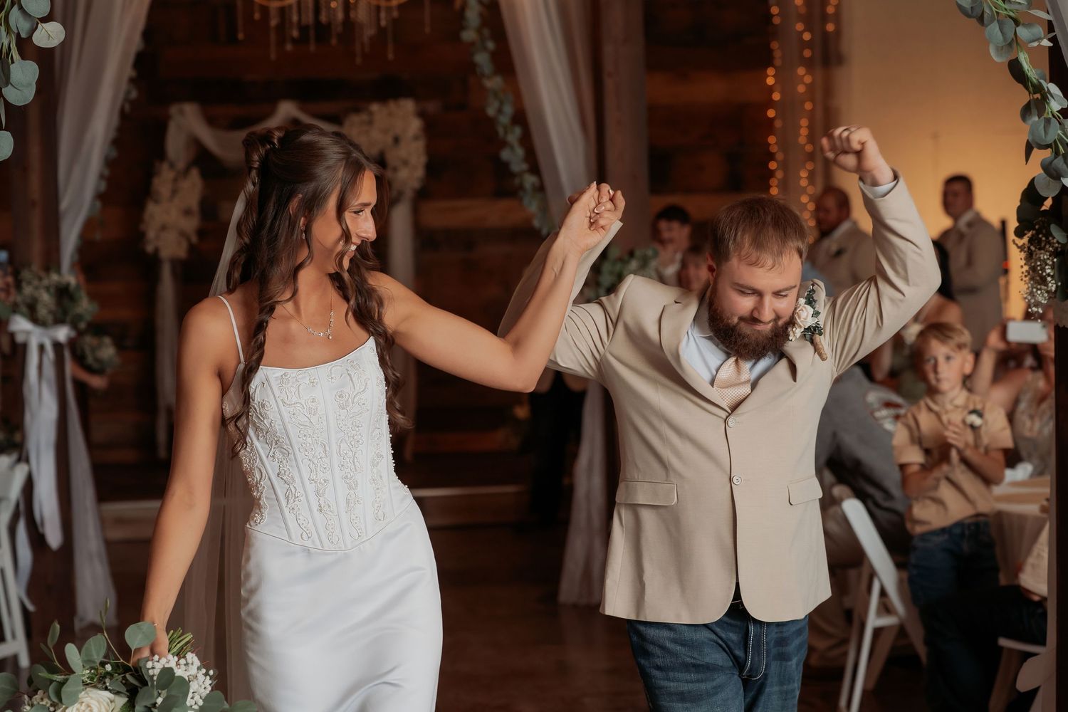 Bride in white gown and groom in tan blazer celebrate their wedding reception entrance with raised hands.