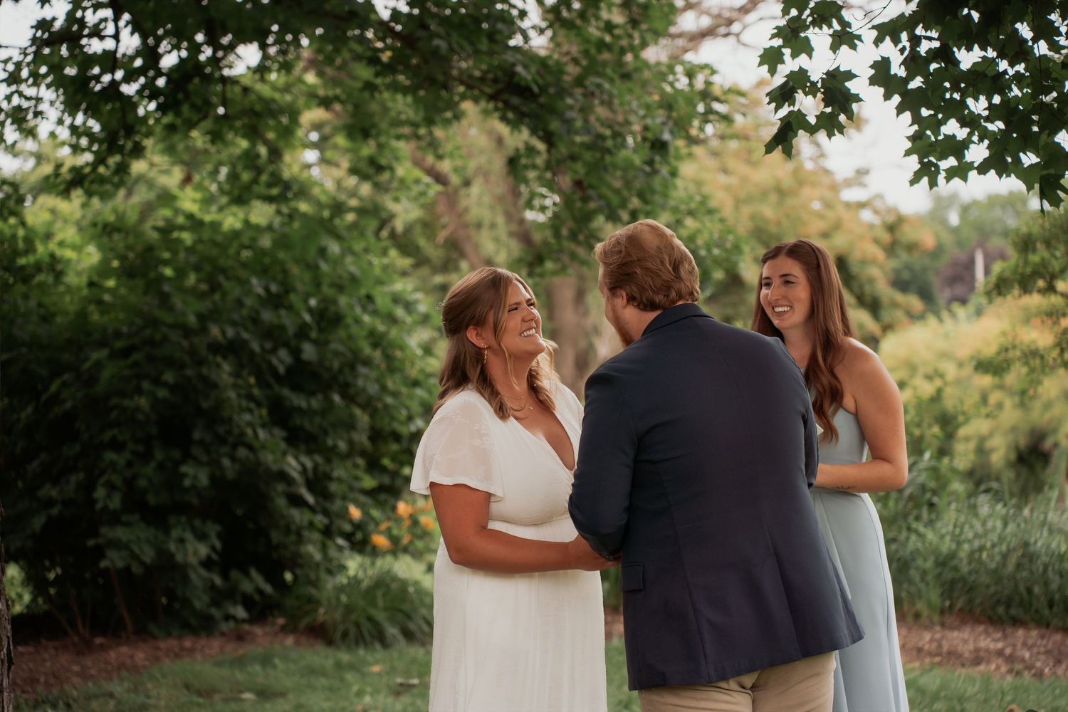Bride and groom exchange vows outdoors with officiant in lush green garden setting during intimate wedding ceremony.