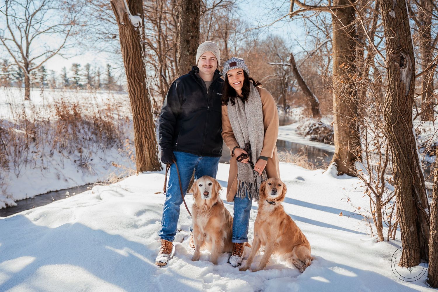 A couple poses with their two golden retrievers on a snowy winter trail.