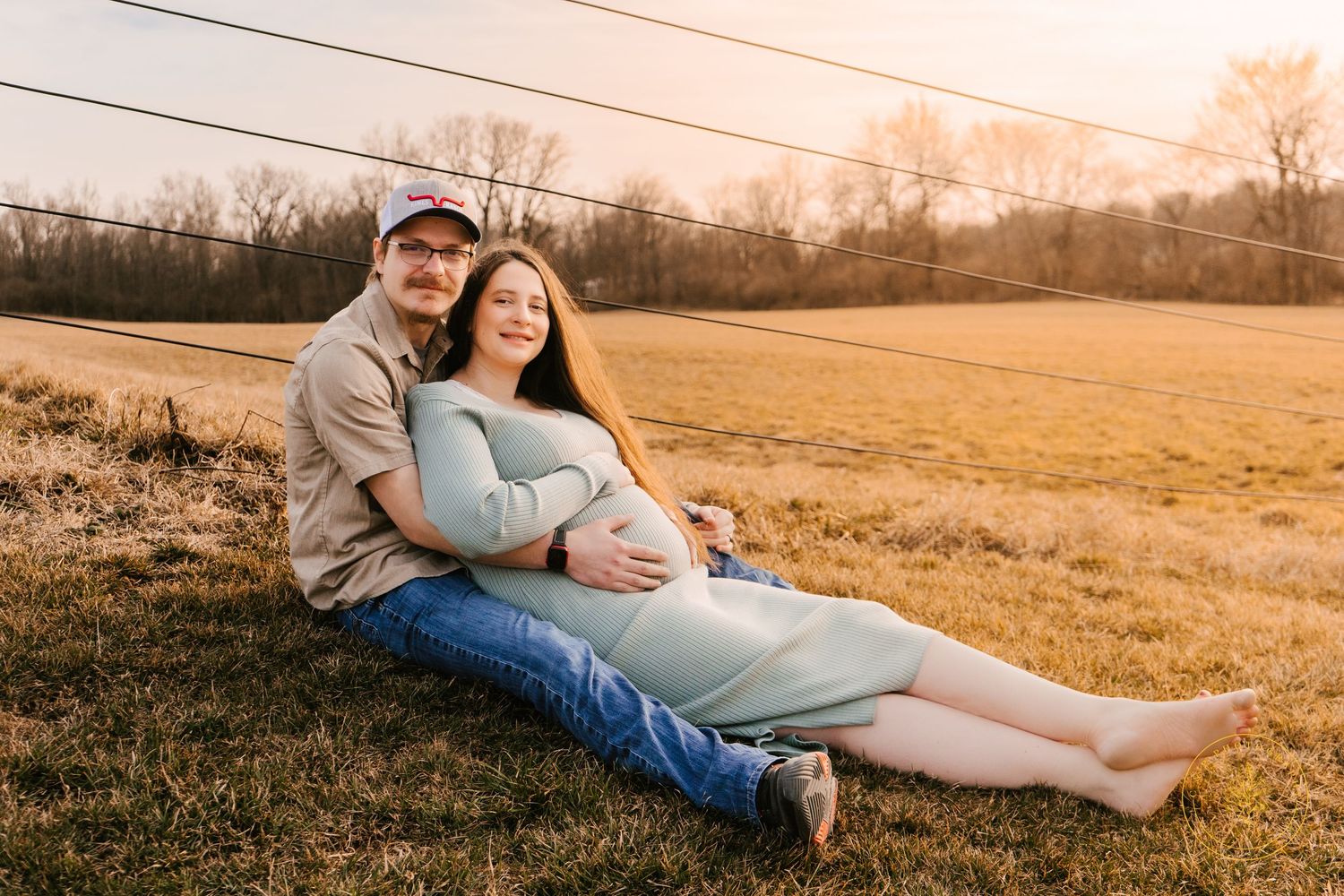 Couple lounging together in a golden field at sunset during maternity photoshoot.
