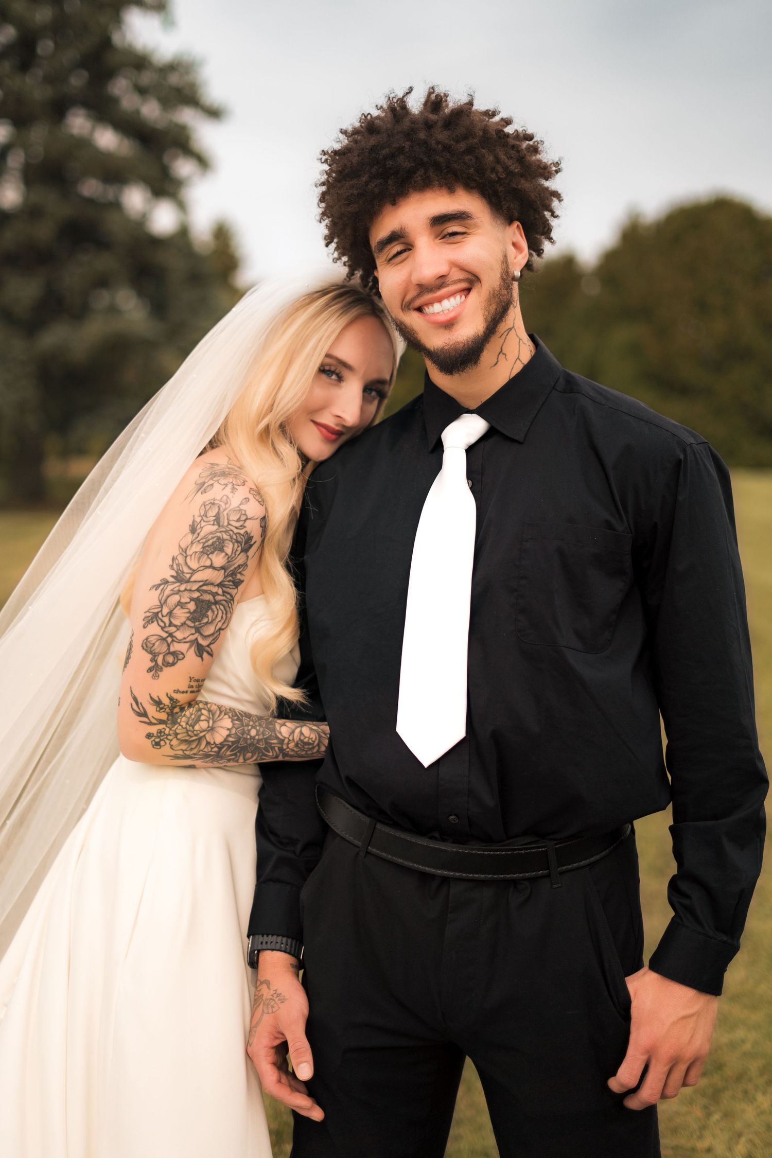 Newlywed couple poses outdoors, bride in white dress with tattoos and groom in black suit.