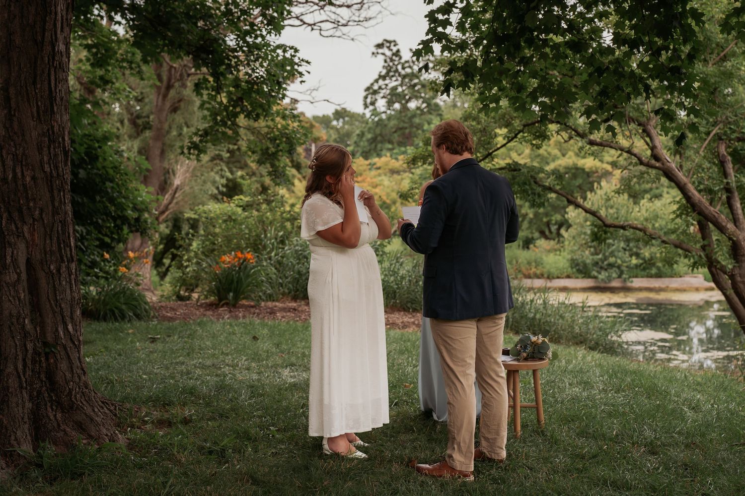 Couple exchanging vows outdoors near a pond surrounded by lush green trees during an intimate garden wedding ceremony.