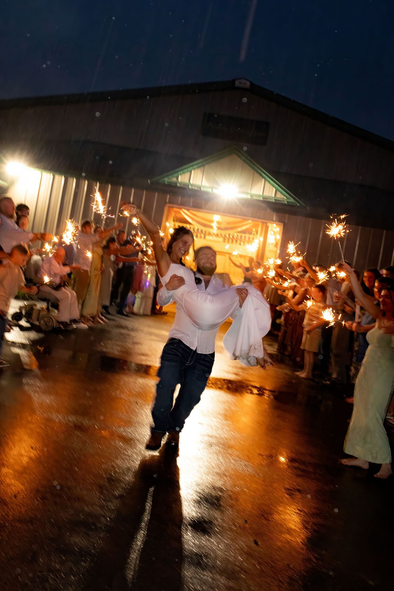 Newlywed couple runs through sparkler tunnel at rainy night wedding reception outside barn venue.