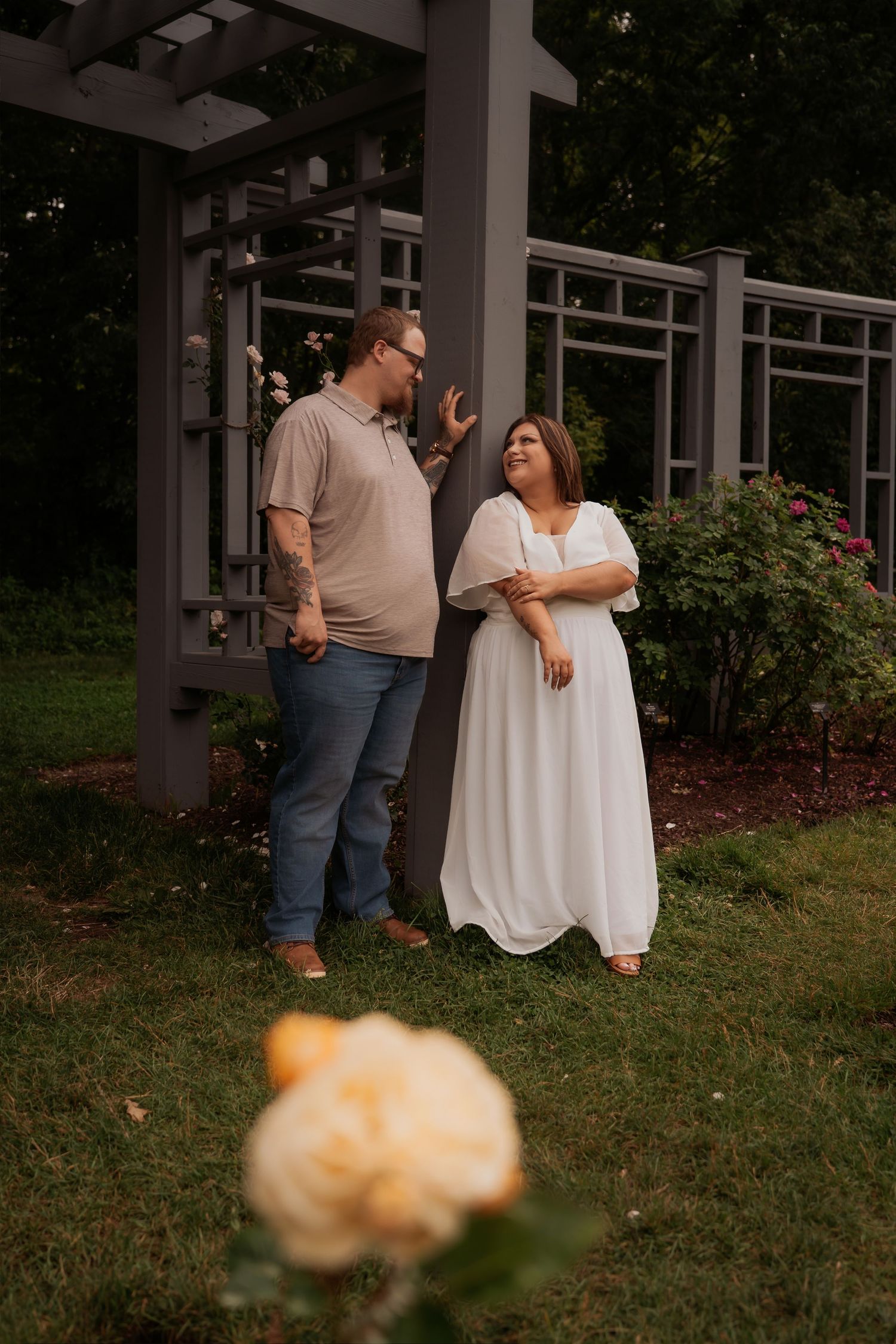 Couple sharing a romantic moment in a garden pergola, woman in white dress and man in casual attire among roses.