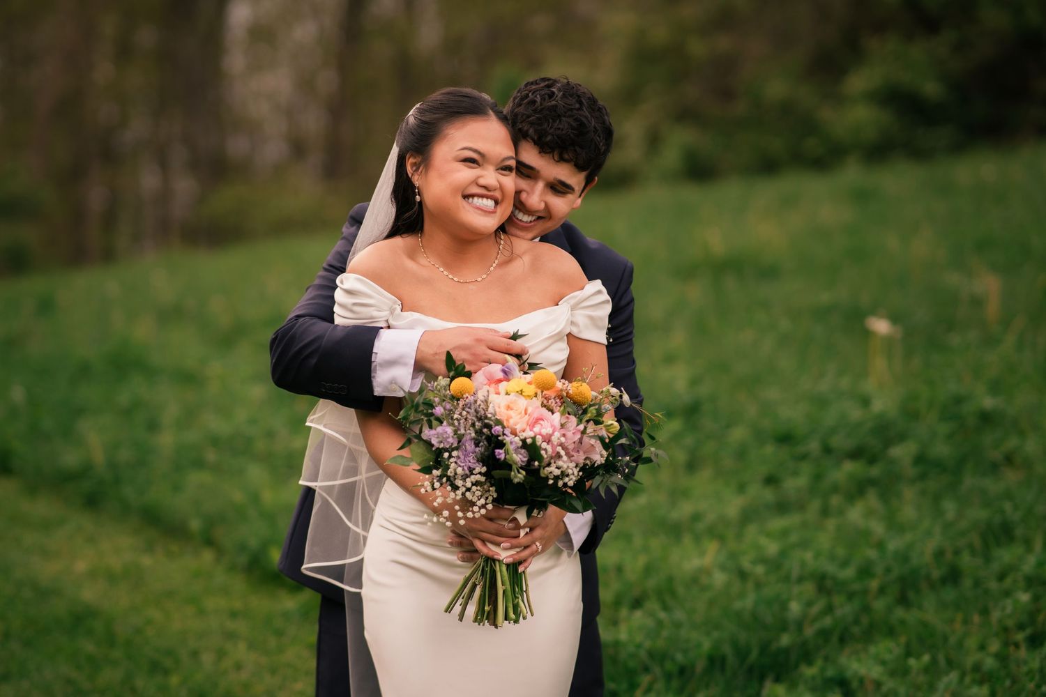 Bride in white gown holding floral bouquet embraced by groom in navy suit on lush green lawn.