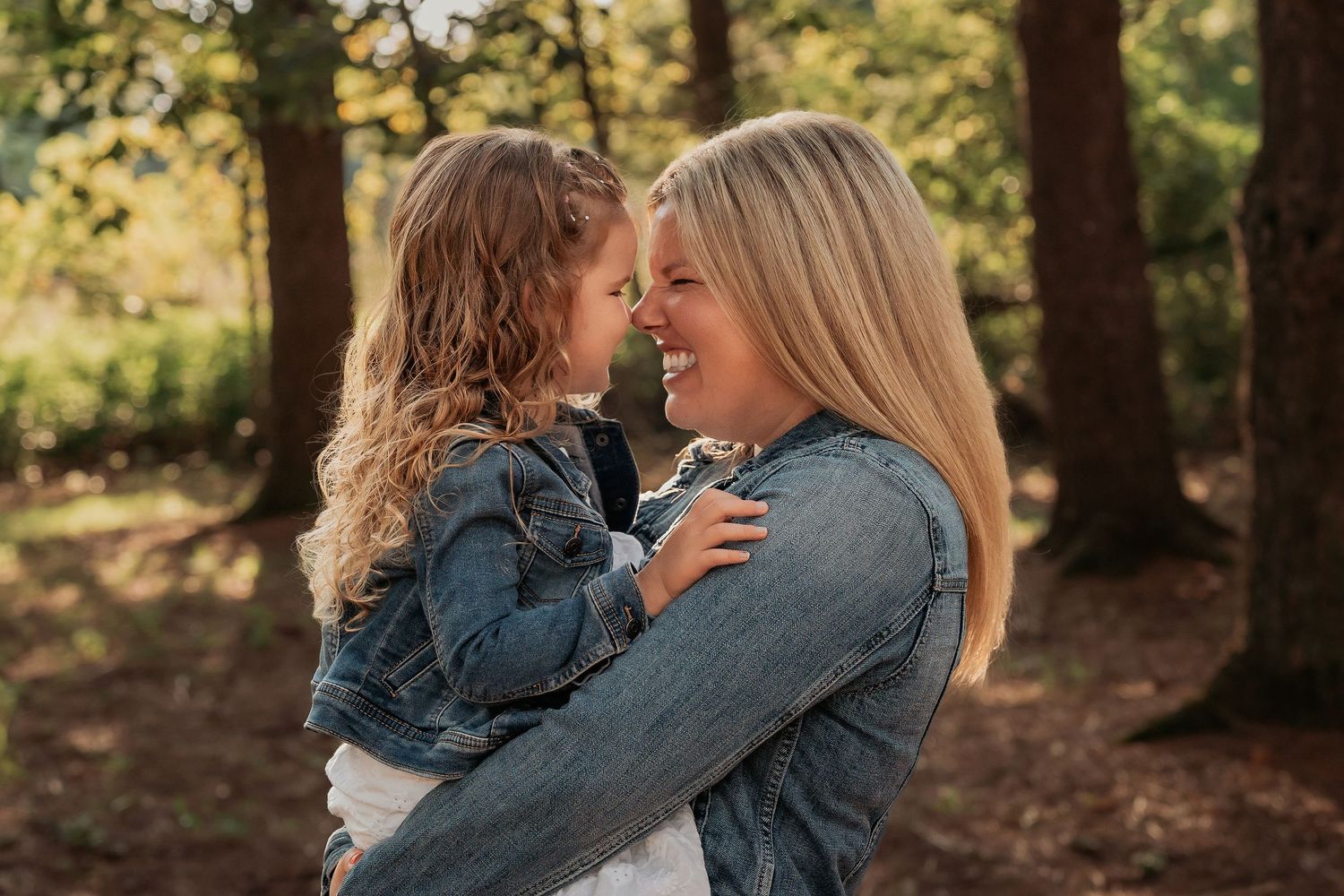 A smiling blonde woman holds a curly-haired girl in a denim jacket outdoors among trees.