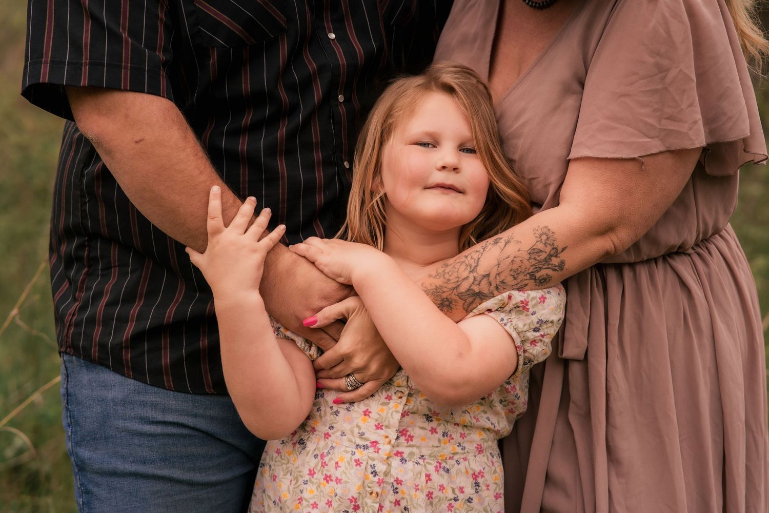 A young girl smiles while being embraced by her parents during an outdoor family photo session in a grassy field.