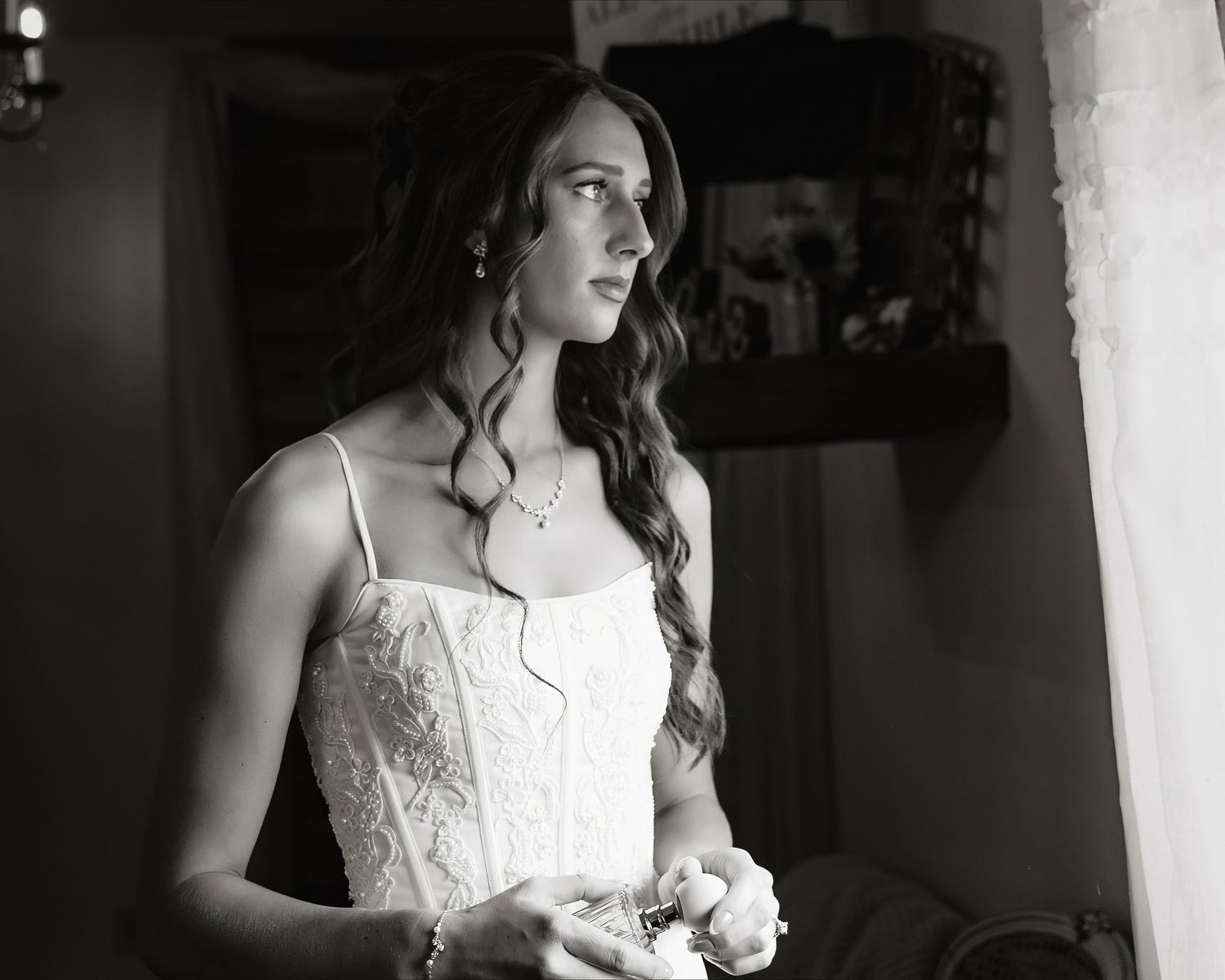 Black and white photo of a bride in a lace gown holding flowers, gazing thoughtfully out a window on her wedding day.