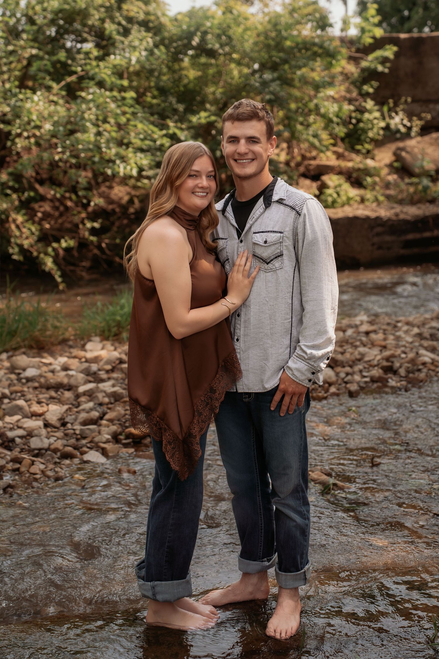 Couple standing barefoot in a shallow creek, smiling, surrounded by lush greenery and rocks.