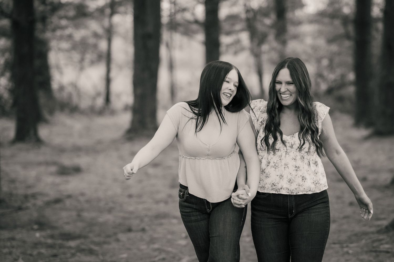Black and white photo of two smiling young women walking hand in hand through a wooded forest path.