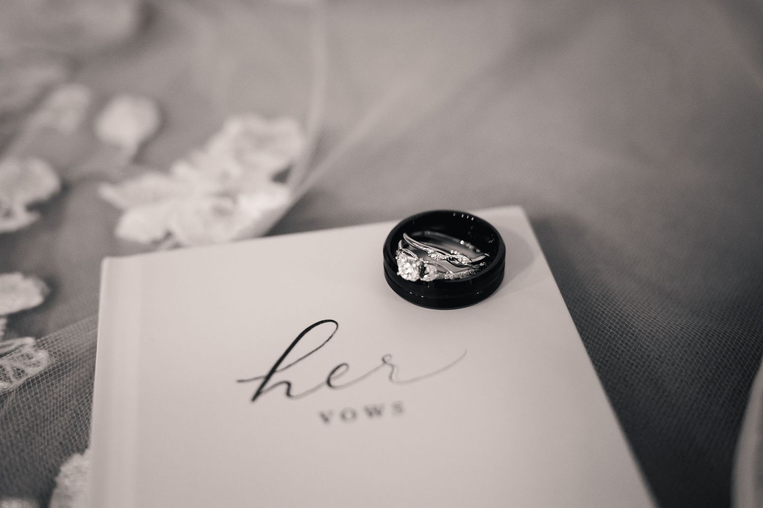 Black and white photo of wedding rings resting on a vow book labeled her vows with a bridal veil in background.