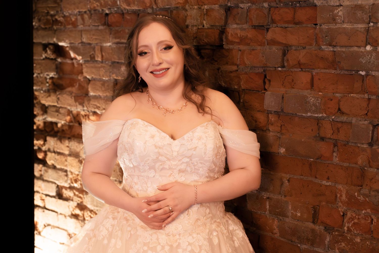 Smiling bride in off-shoulder lace ball gown posing against rustic brick wall in elegant wedding portrait.