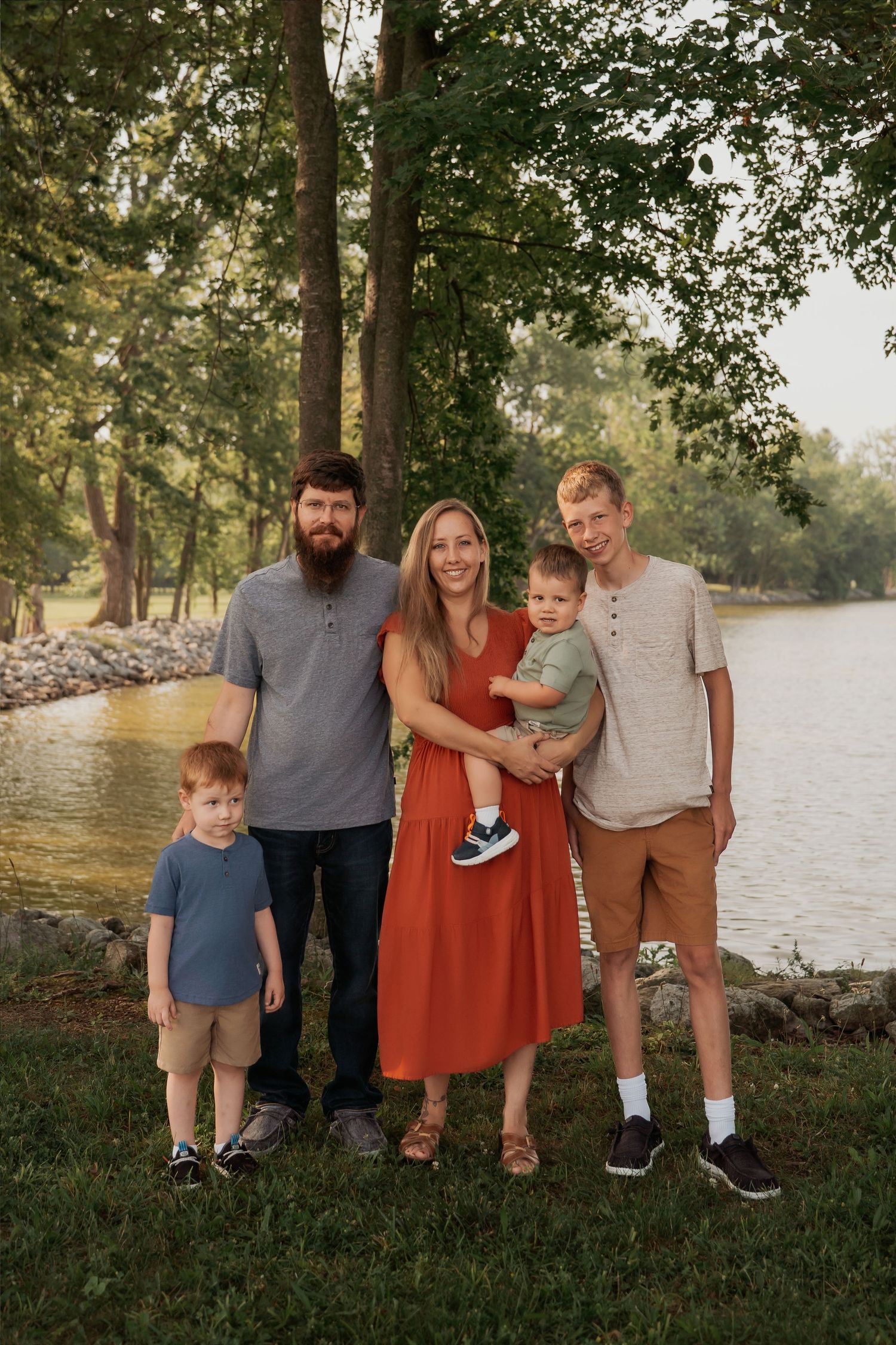 Family of five posing outdoors near a lake surrounded by trees during golden hour.