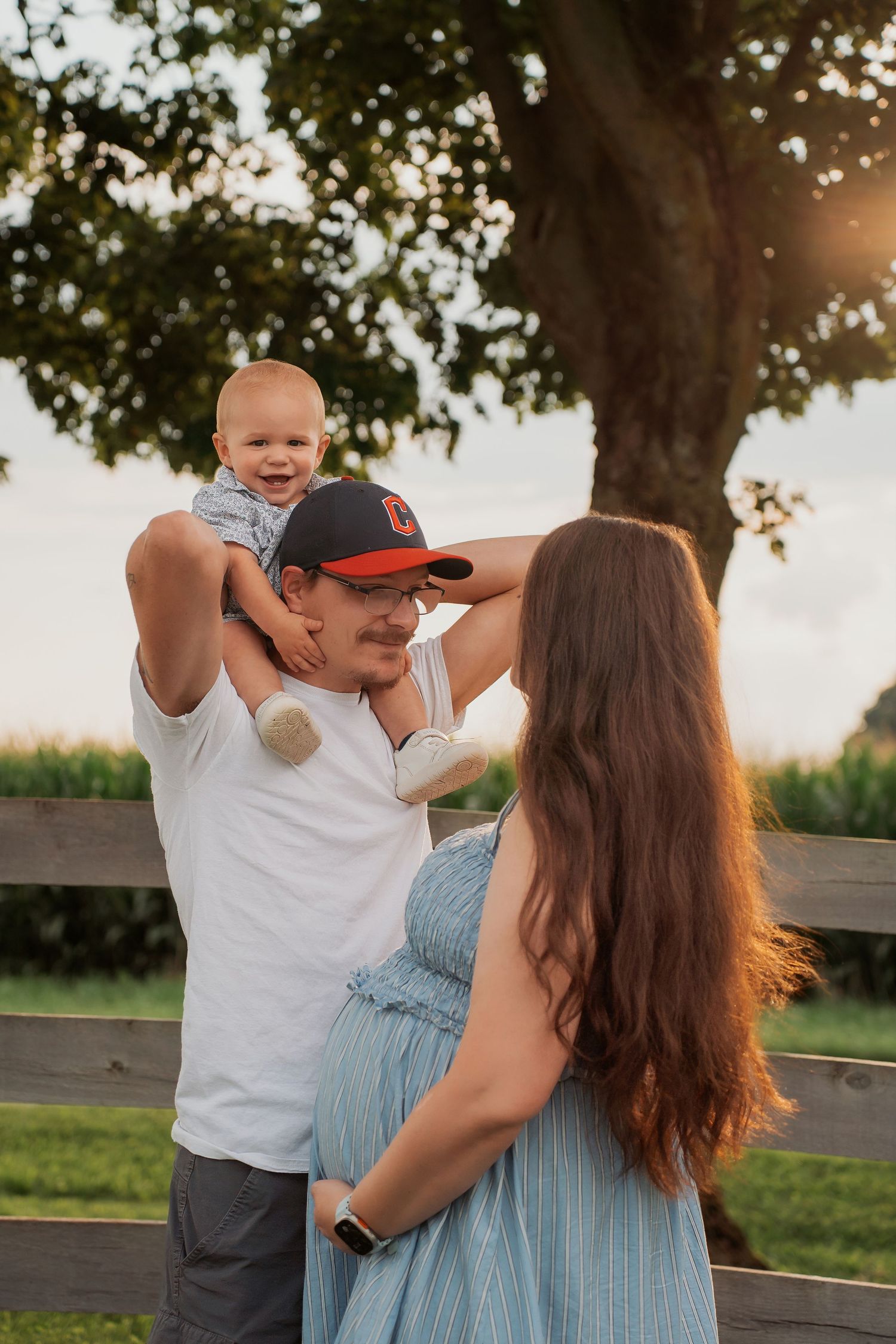 A happy family outdoors at golden hour, father lifting smiling baby while pregnant mother watches near a wooden fence.