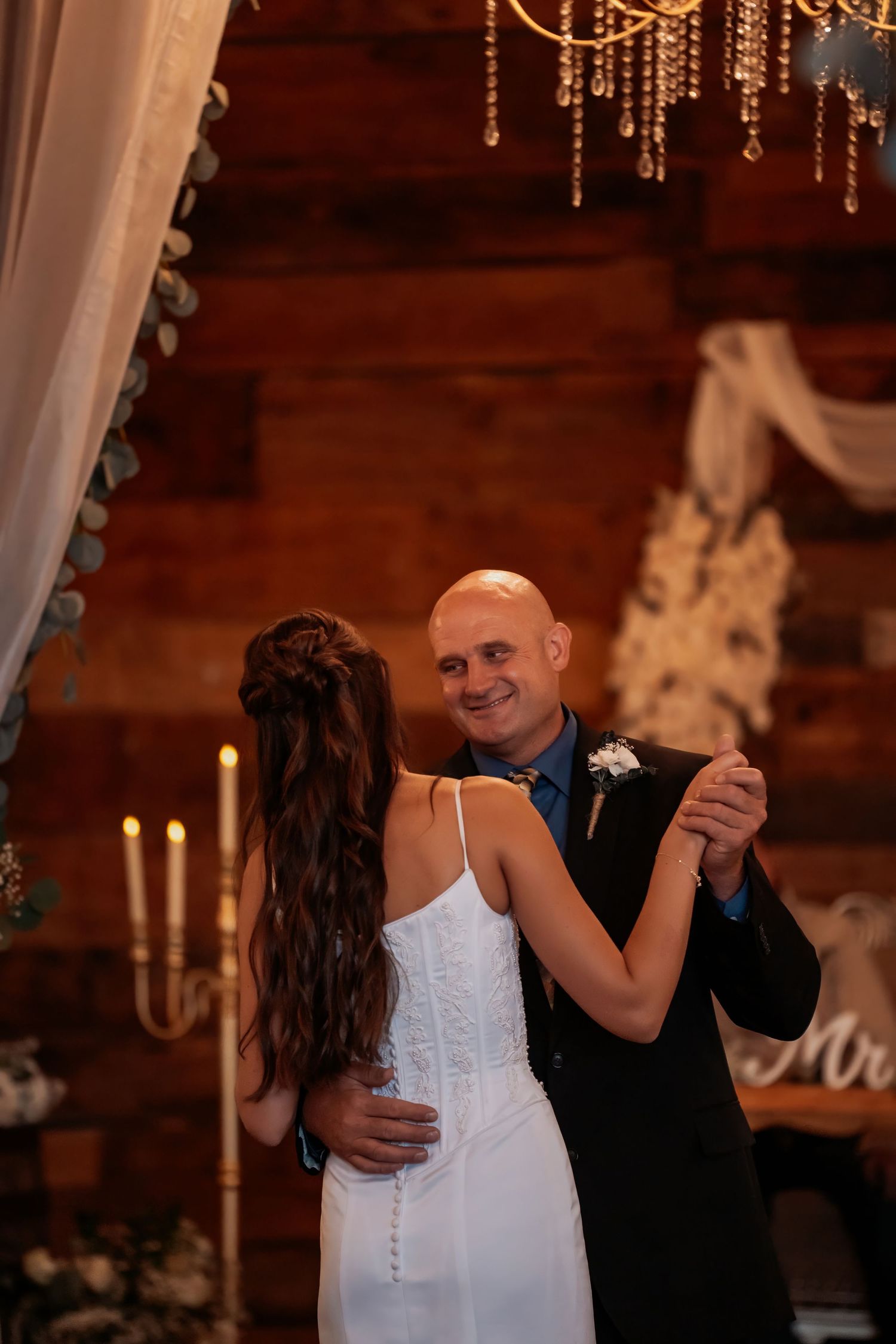 Bride and groom share their first dance in a rustic barn wedding venue with warm candlelight and elegant chandelier.