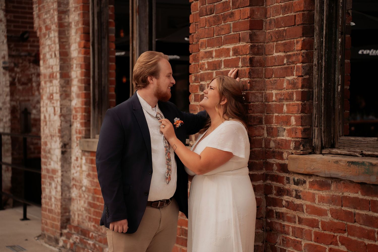 Couple in wedding attire sharing a romantic moment against a rustic brick wall outdoors.